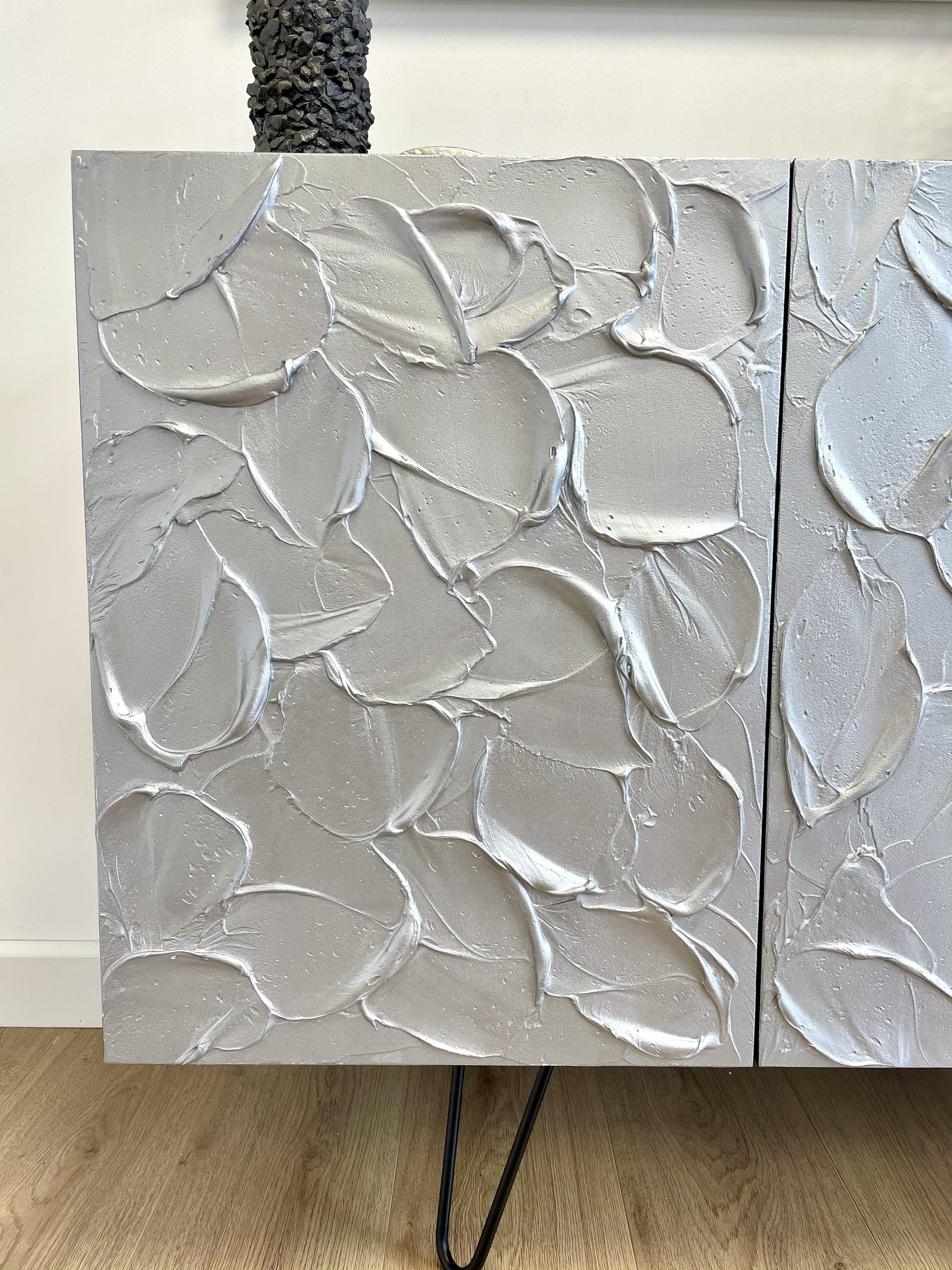 Close-up of a textured art cabinet with a modern design. The surface showcases thick, overlapping strokes of gray material in a leaf-like pattern. This textured piece stands on black metal legs.
