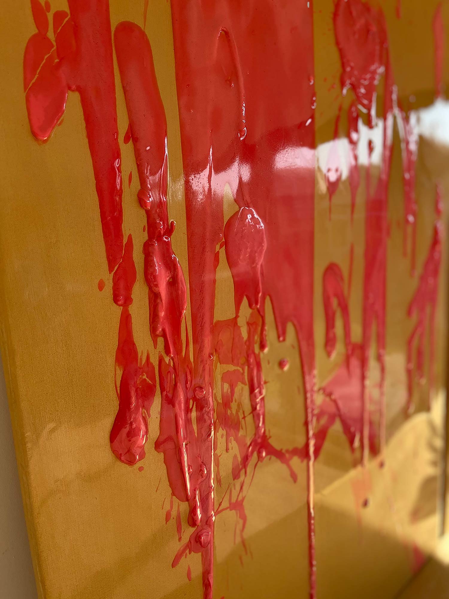 Close-up of a canvas with bright red, glossy paint dripping down a yellow background.