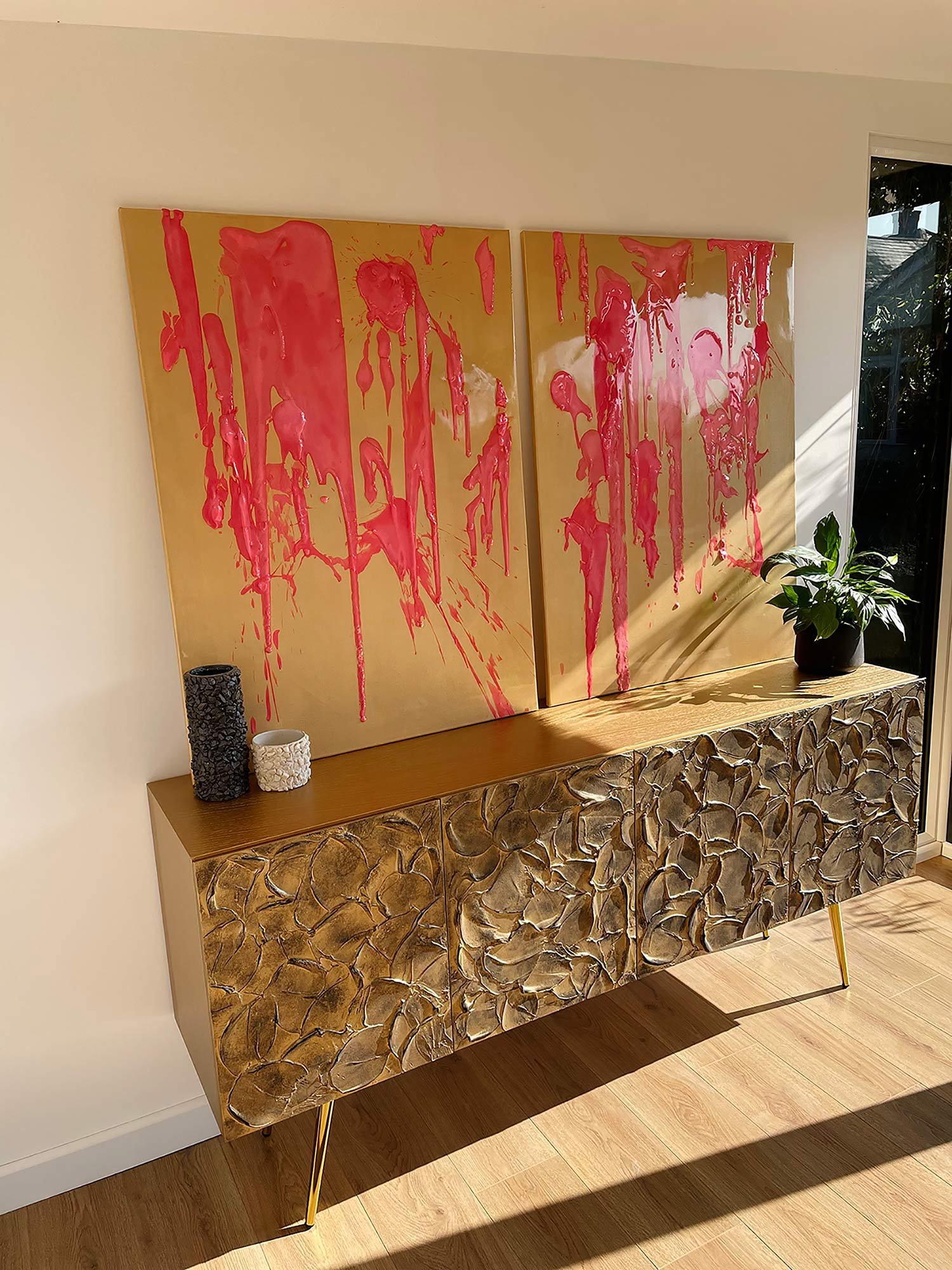 Gold tetured sideboard. On top, there are two large gold panels with bold pink paint splatters.