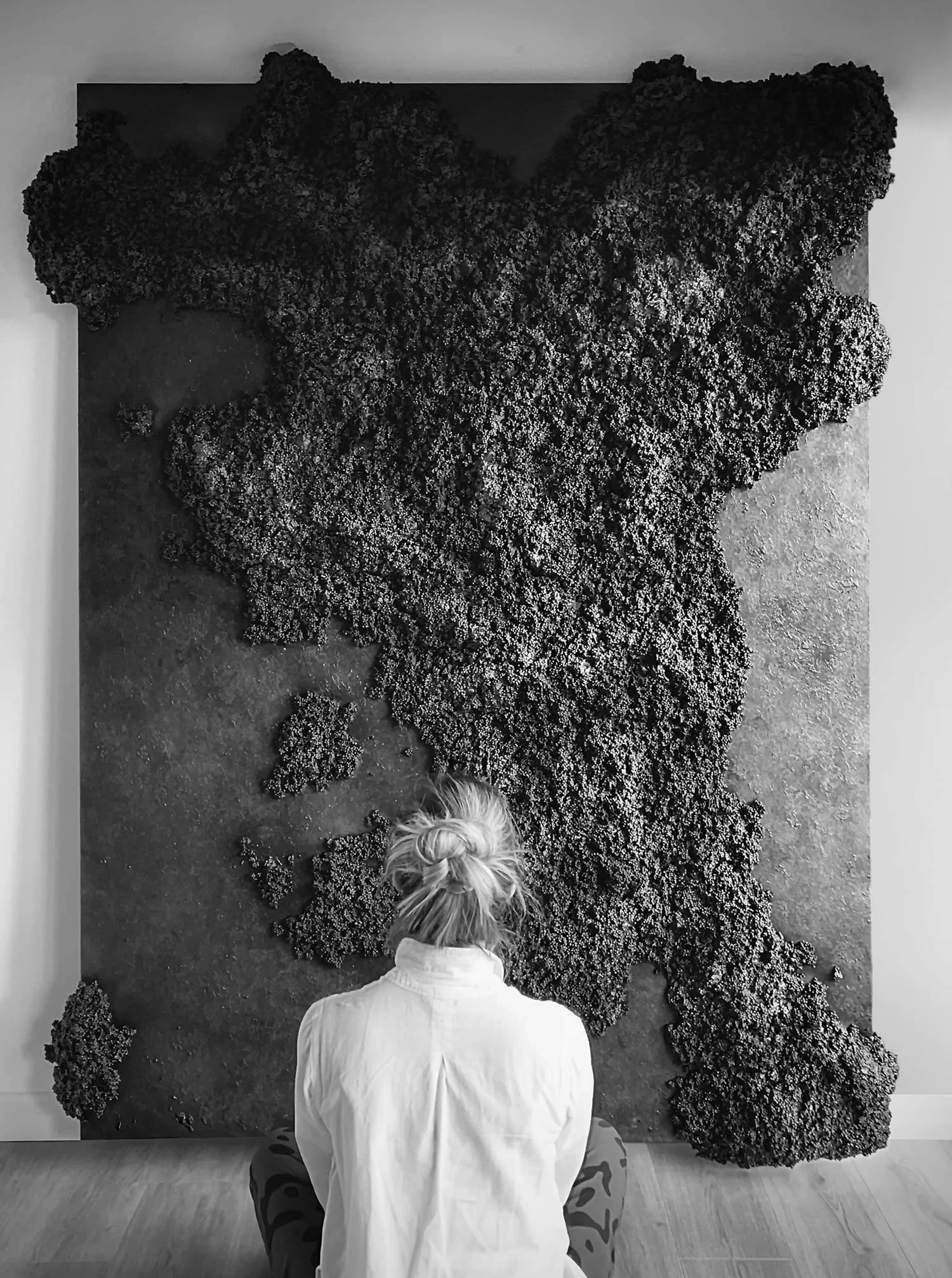Large, black, abstract wall art made from recycled materials hanging on the wall, with woman sitting in front of it.