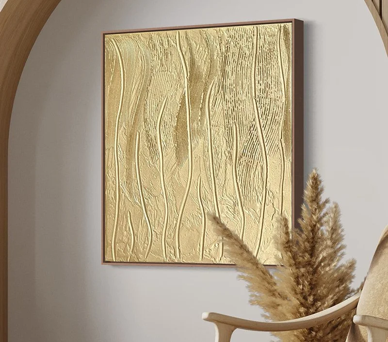 A modern living room corner featuring a large abstract gold textured artwork on a gray wall, a beige armchair, a potted pampas grass plant, and a small table with books.
