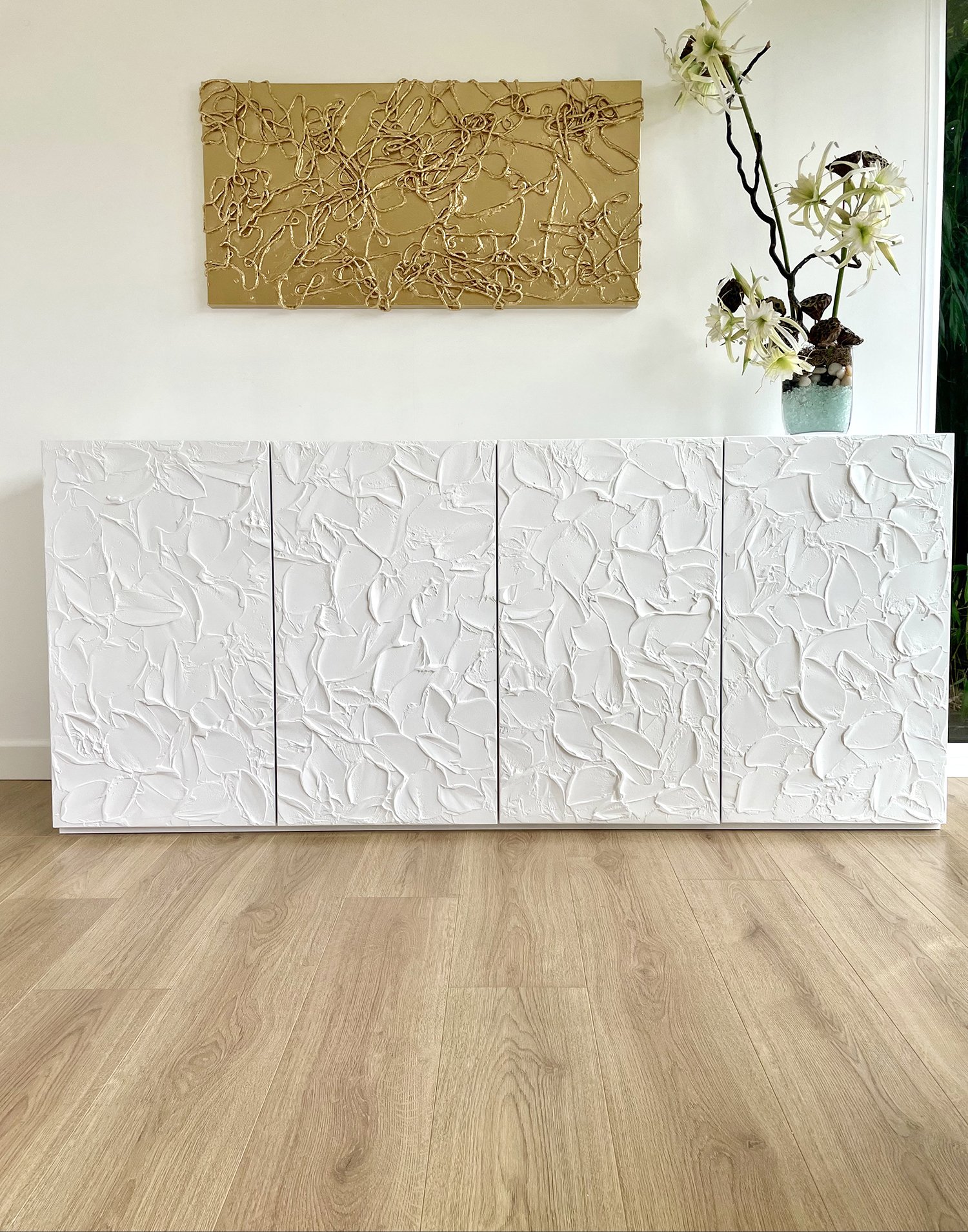 White textured cabinet in a modern room with light wooden flooring. Above it, a gold abstract wall art piece. A vase with white and purple flowers sits on the cabinet’s corner.
