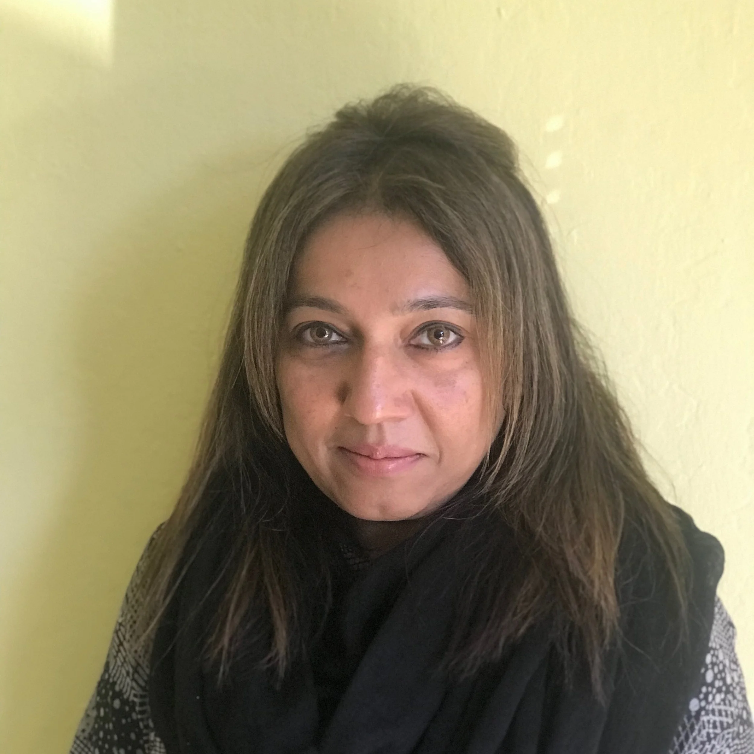 Image of Shakila. She is smiling and standing on front of a yellow background. She is wearing a black and white shirt with a black scarf.