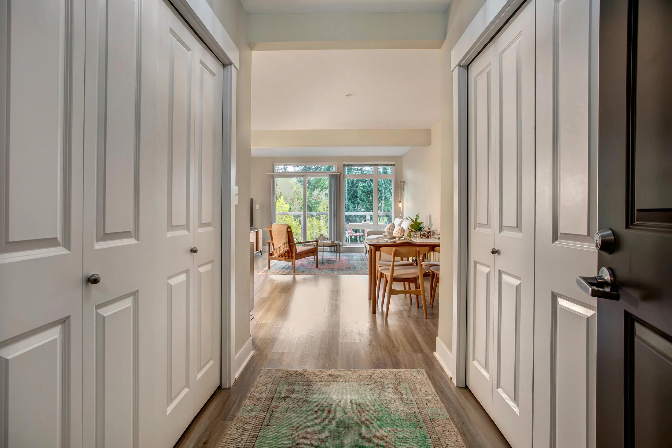 View from a doorway into a living room with large windows, wooden flooring, and furniture including chairs and a table, with sunlight and greenery visible through the windows.