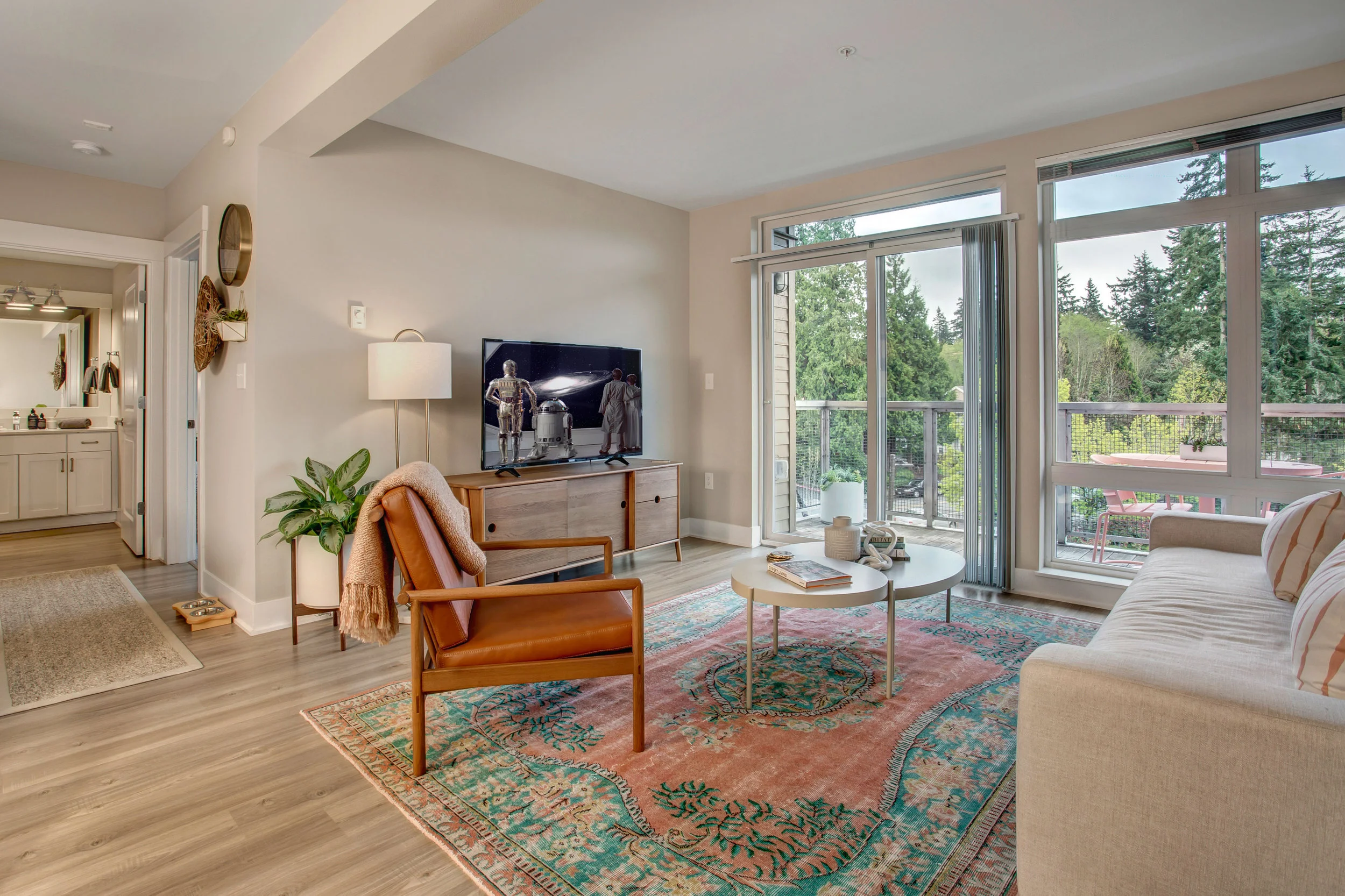 Living room with wooden furniture, a colorful area rug, a beige sofa, a side table, a flat-screen TV, and large windows showing an outdoor balcony with pink chairs and green trees in the background.
