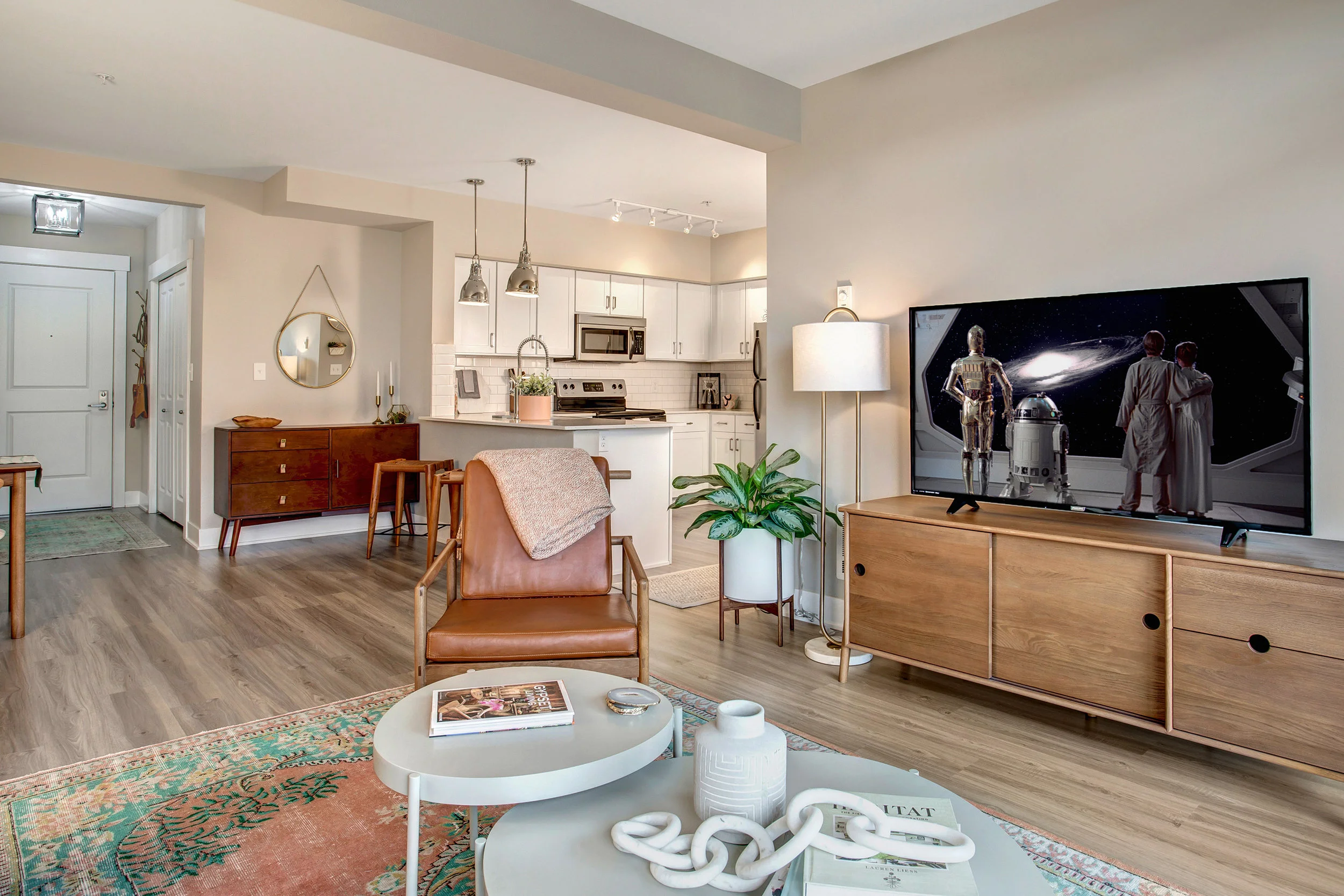 Living room with a wooden TV stand, a large flat-screen TV showing characters in space, a lamp, a green potted plant, a chair with a blanket, a coffee table with magazines and decor, and a view of an open-concept kitchen with white cabinets and stainless steel appliances.