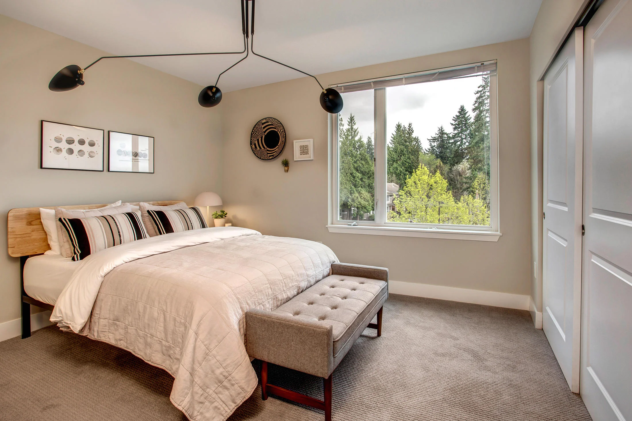 A bedroom with a bed, striped pillows, a small side table with a lamp, wall art, a window showing trees outside, and a closet with sliding doors.