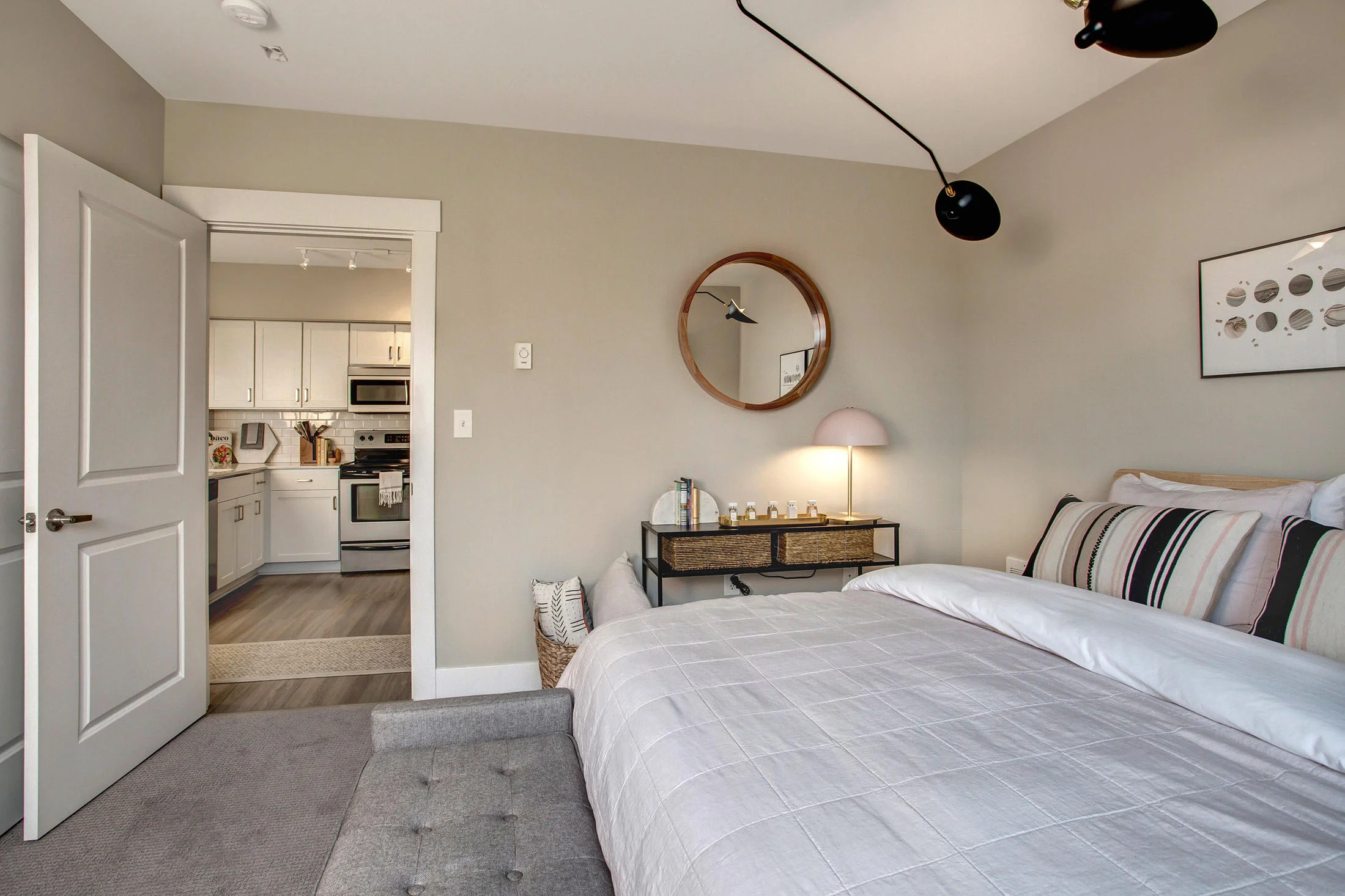 A cozy bedroom with a bed featuring striped pillows and a white quilt, a black metal console table with baskets, a small lamp, and framed art on the wall. An opening reveals a kitchen with white cabinets and appliances.