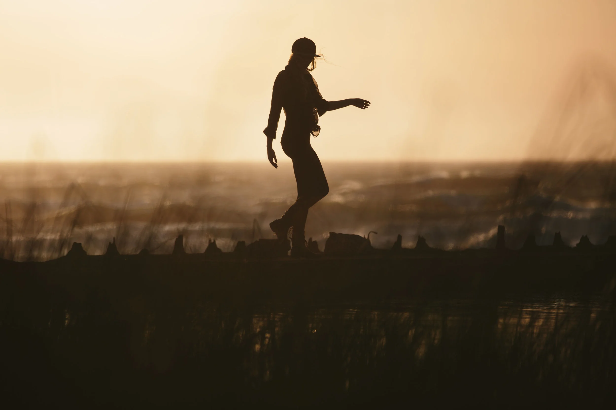 Silhouette of a person wearing a cap walking on a beach at sunset or sunrise with the ocean in the background.