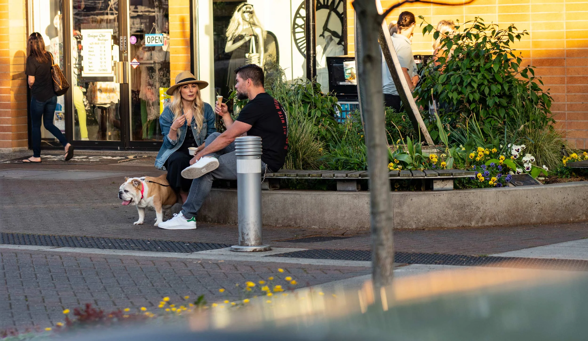 Two people sitting on a bench outside a storefront, having a conversation. The woman is wearing a hat and the man is holding an ice cream cone. A bulldog with a pink collar is on the ground next to them. There are people walking by, and a display win
