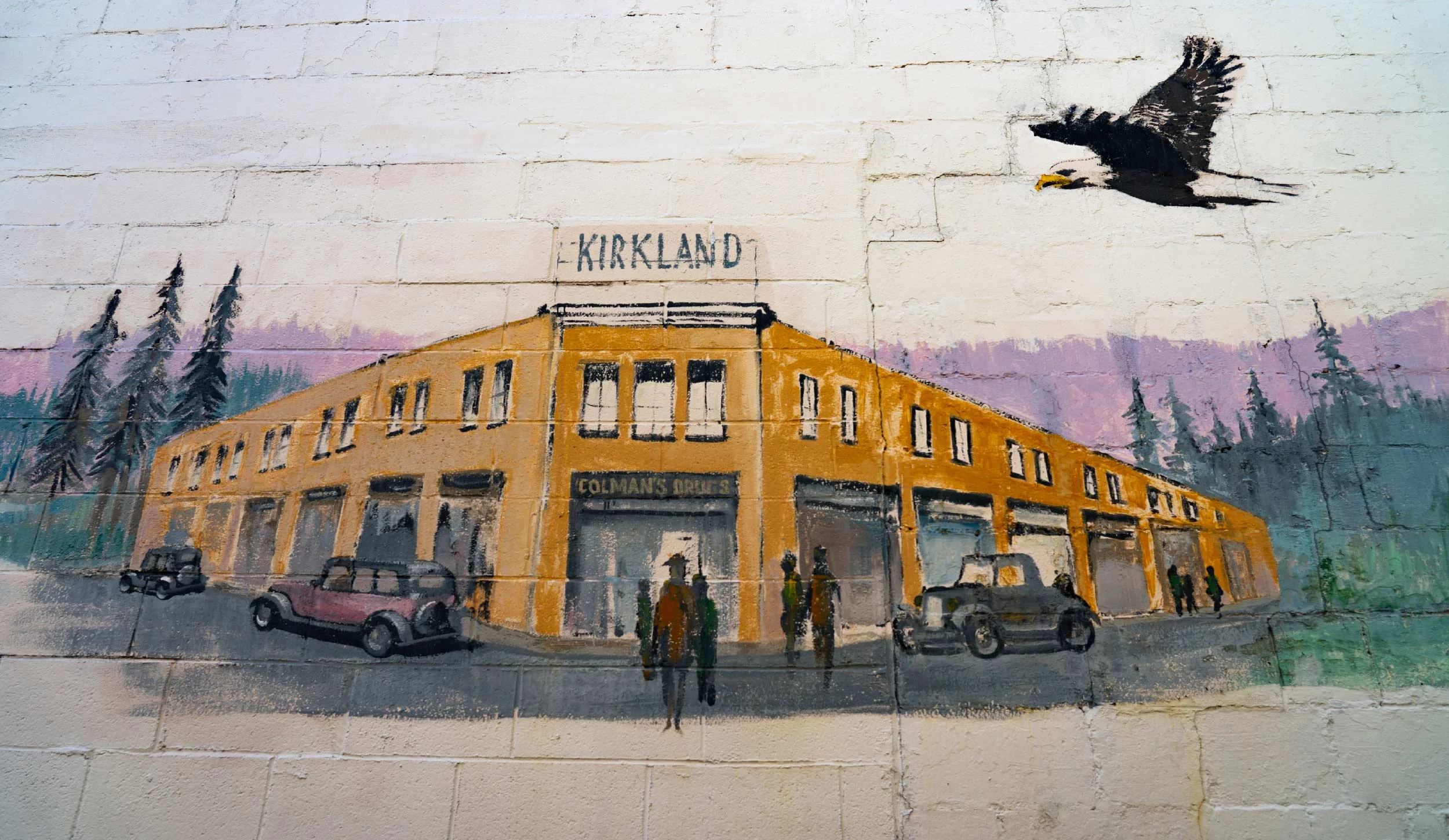 A mural on a brick wall depicts a yellow building with a sign that reads 'Kirkland' and a street scene with vintage cars and people. A large eagle is flying above the building.