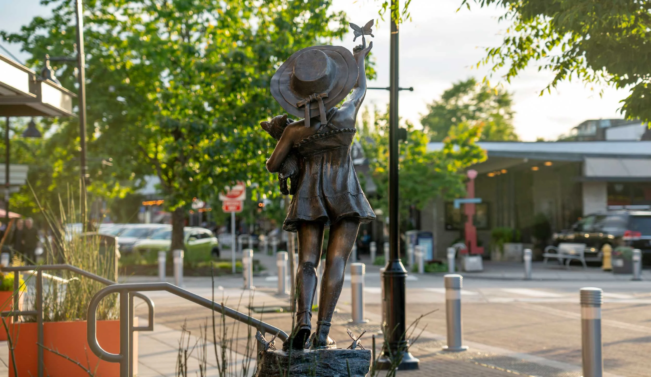 Bronze statue of a girl holding a hat, standing on a sidewalk in an urban area with trees, lampposts, and parked cars in the background during sunset.