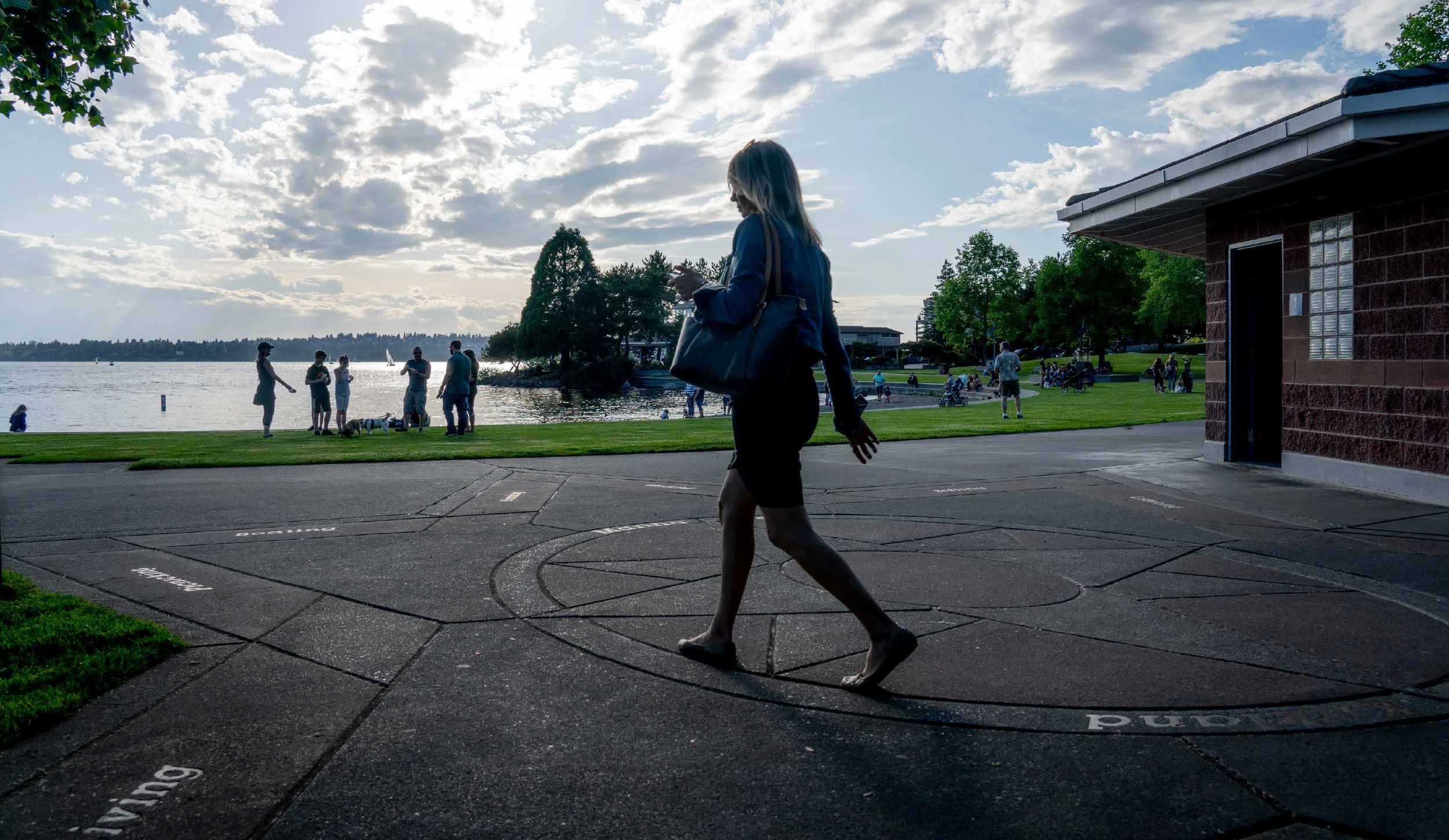 A woman walking near a lakeside park, with people enjoying the outdoor area, on a partly cloudy day with a view of the water and sailboats in the background.