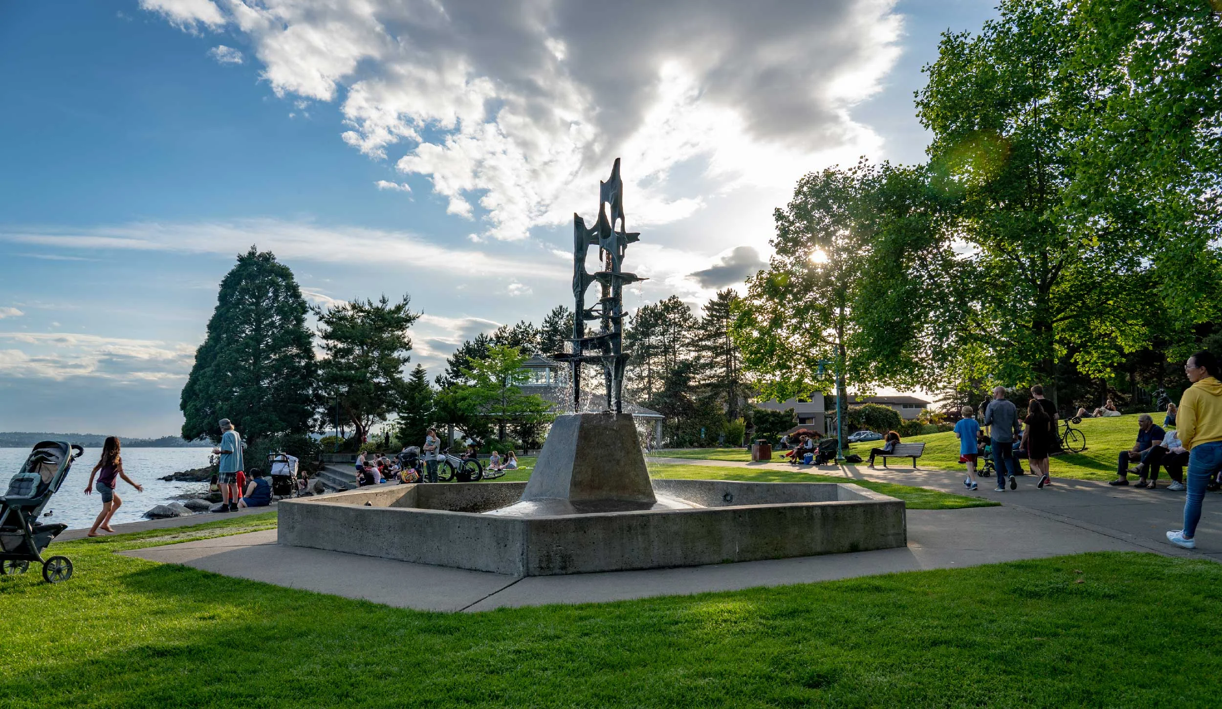 People gathered in a park near a large water body with a modern abstract sculpture fountain, trees, and a partly cloudy sky with the sun setting.