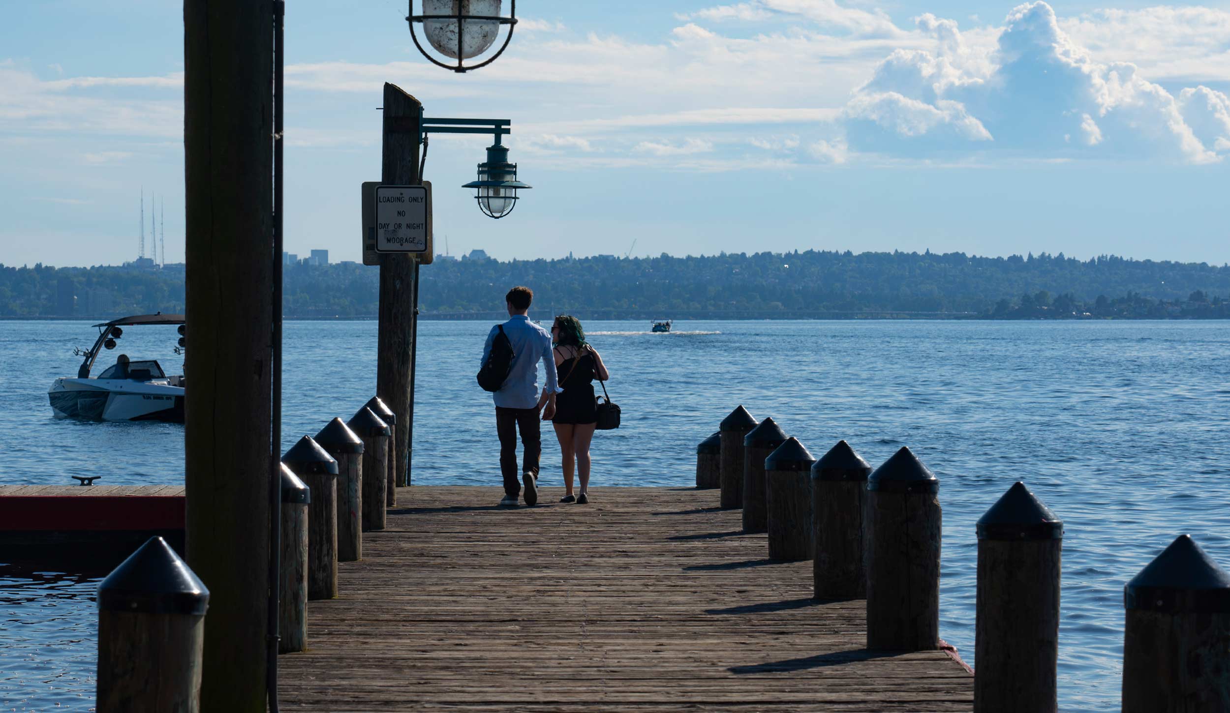 A couple walking on a wooden dock by a large body of water, with a boat and distant shoreline visible in the background, under a partly cloudy sky.