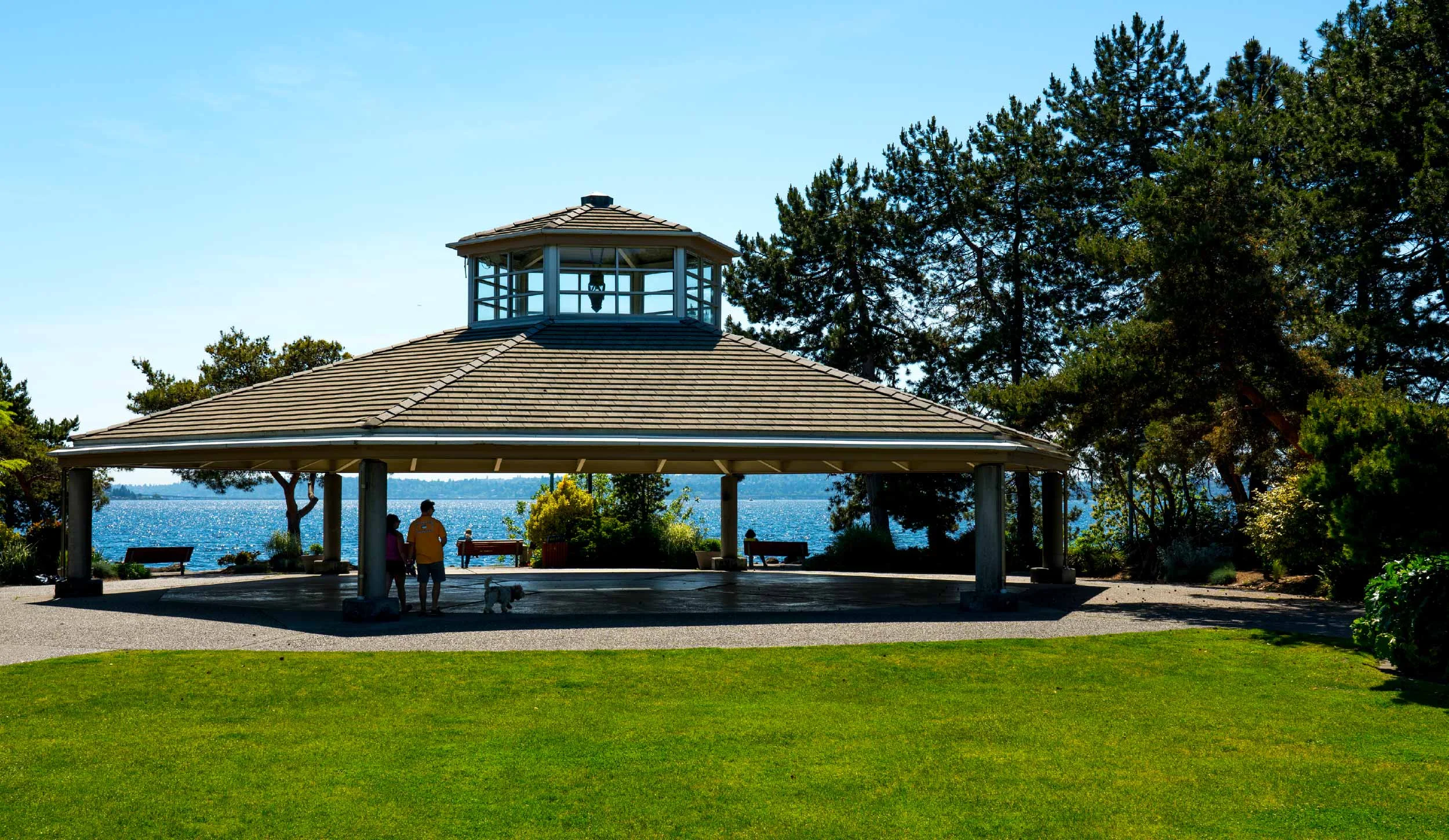 A pavilion with a shingled roof and a lantern in the top window overlooks a lake, surrounded by trees and green grass, with a few people and a dog underneath.
