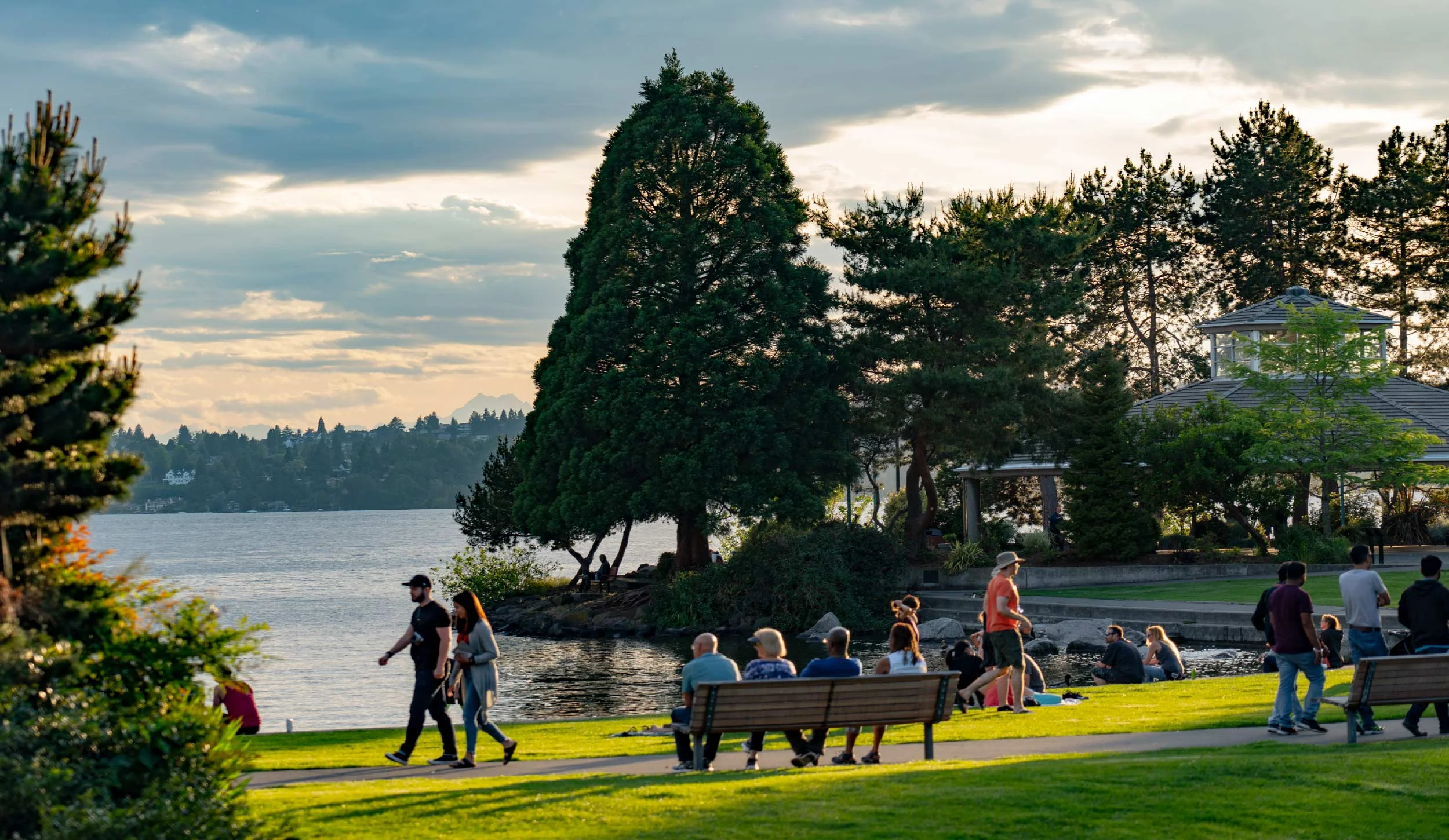 People enjoying a park by the water, with trees and a gazebo, as the sun sets.