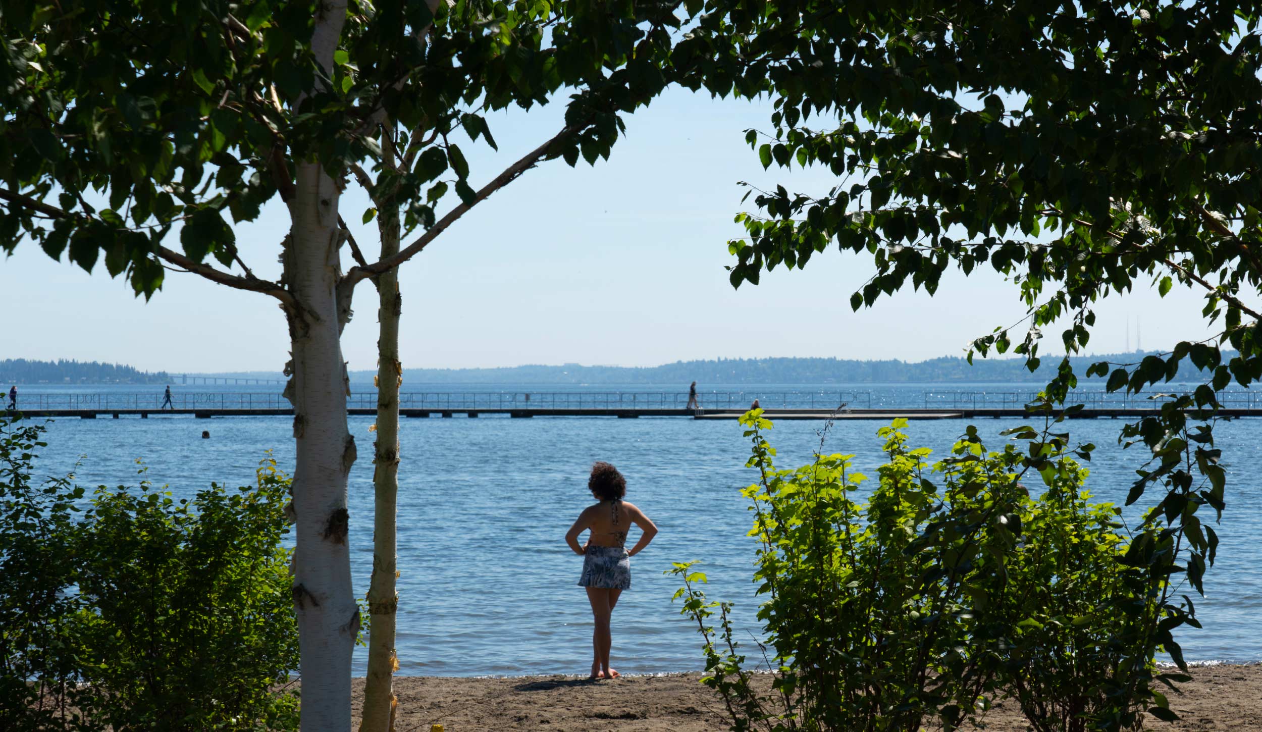 A young woman stands on a sandy beach facing a large body of water, framed by green foliage and trees, with a pier and people walking in the distance across the water.