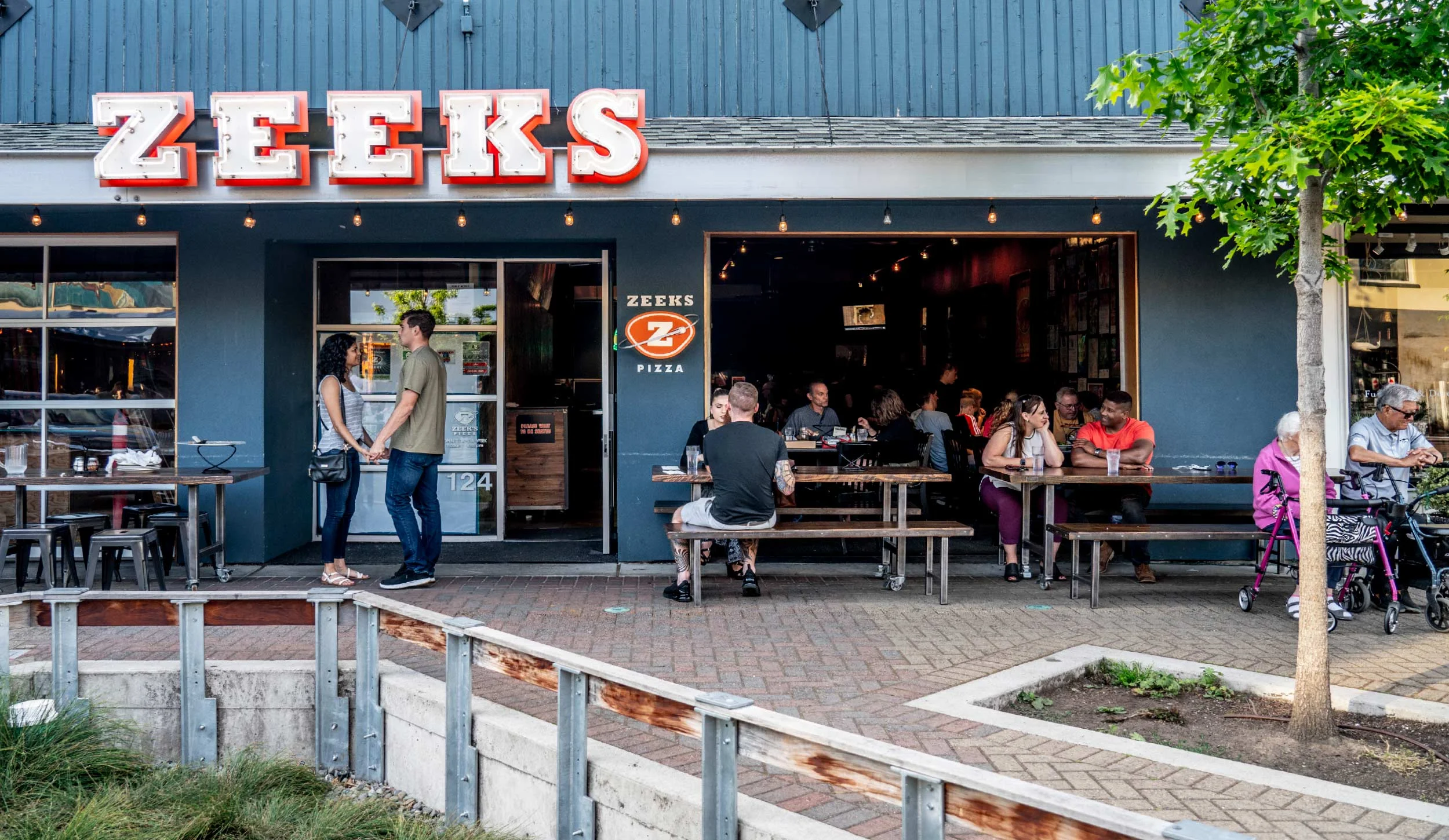 Exterior of a restaurant named Zeeks with customers sitting at outdoor picnic tables and people standing and talking in front of the entrance. There is a leafy tree on the right and a small landscaped area in the foreground.