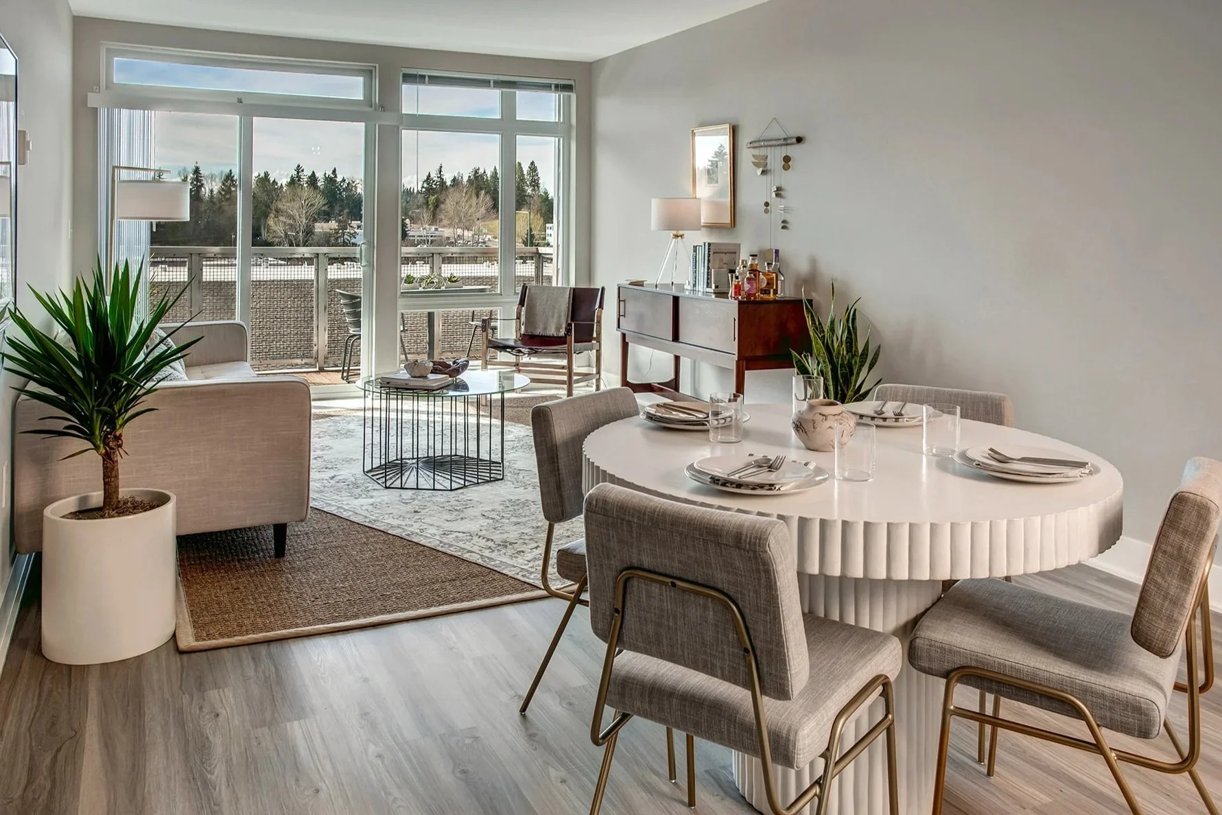 Living and dining area with large windows, houseplants, a white round table with place settings, beige and gray chairs, a glass-top coffee table, a sideboard with decor and liquor bottles, a smaller seating area with a sofa and armchair, and a balcony with outdoor furniture.
