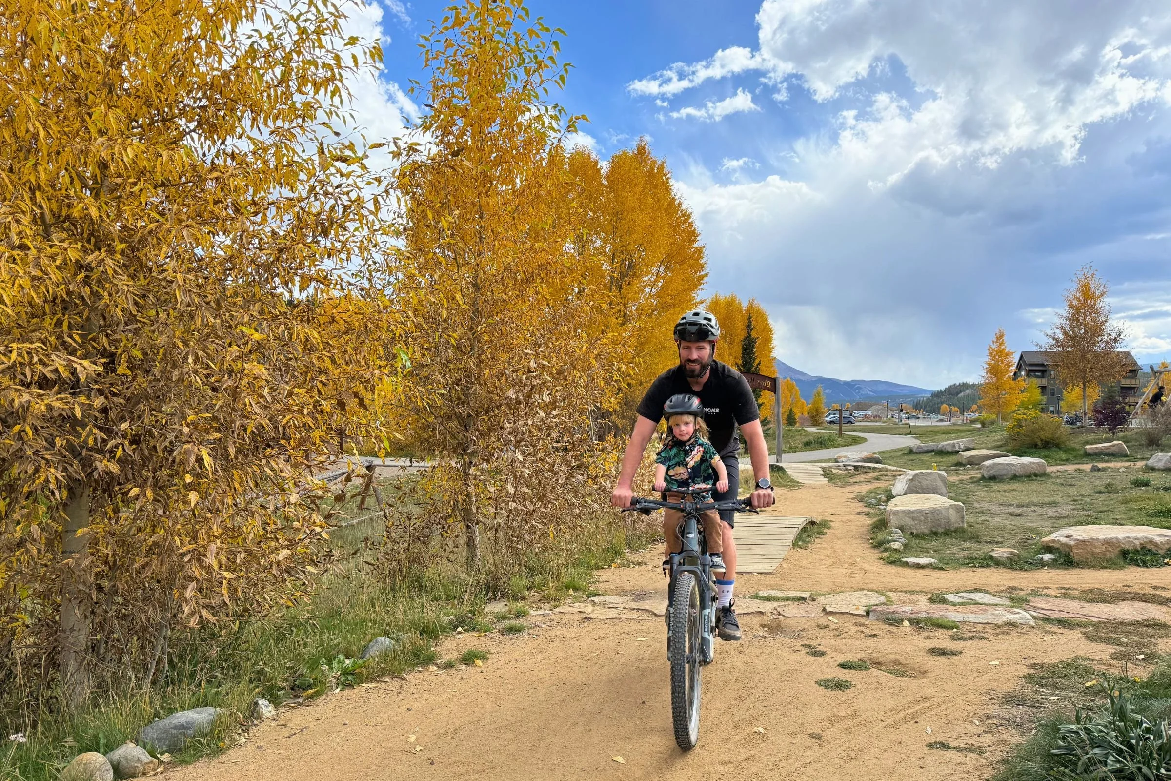 Adult riding a bicycle along a dirt path with a child in the Kids Ride Shotgun Shotgun Pro Evo, with trees turning yellow in the background.