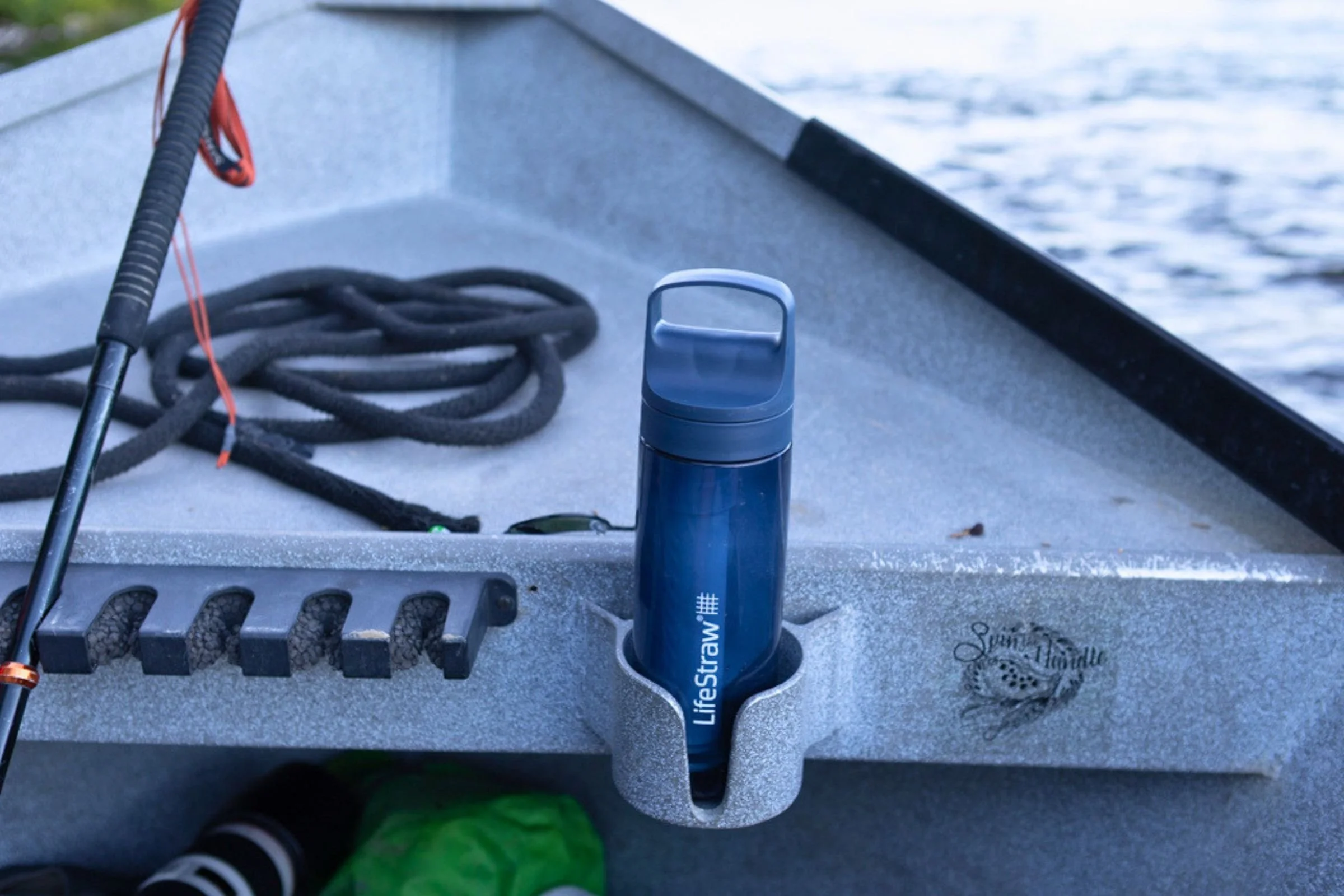 LifeStraw Go Series Travel Water Bottle in a dark blue finish, secured upright in a boat holder beside coiled rope and a fishing rod, with open water visible beyond.