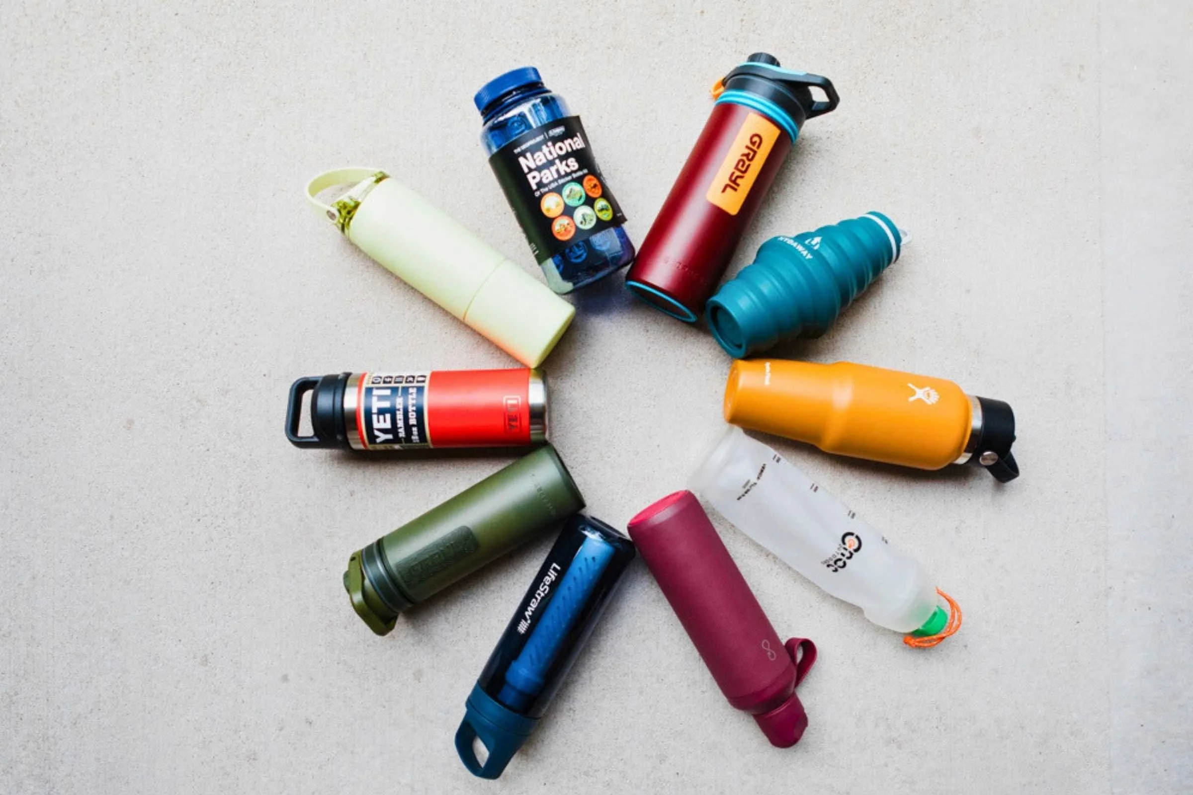 Six travel water bottles laid flat on a neutral surface, including rigid, collapsible, and filter models. Photo courtesy Karthika Gupta