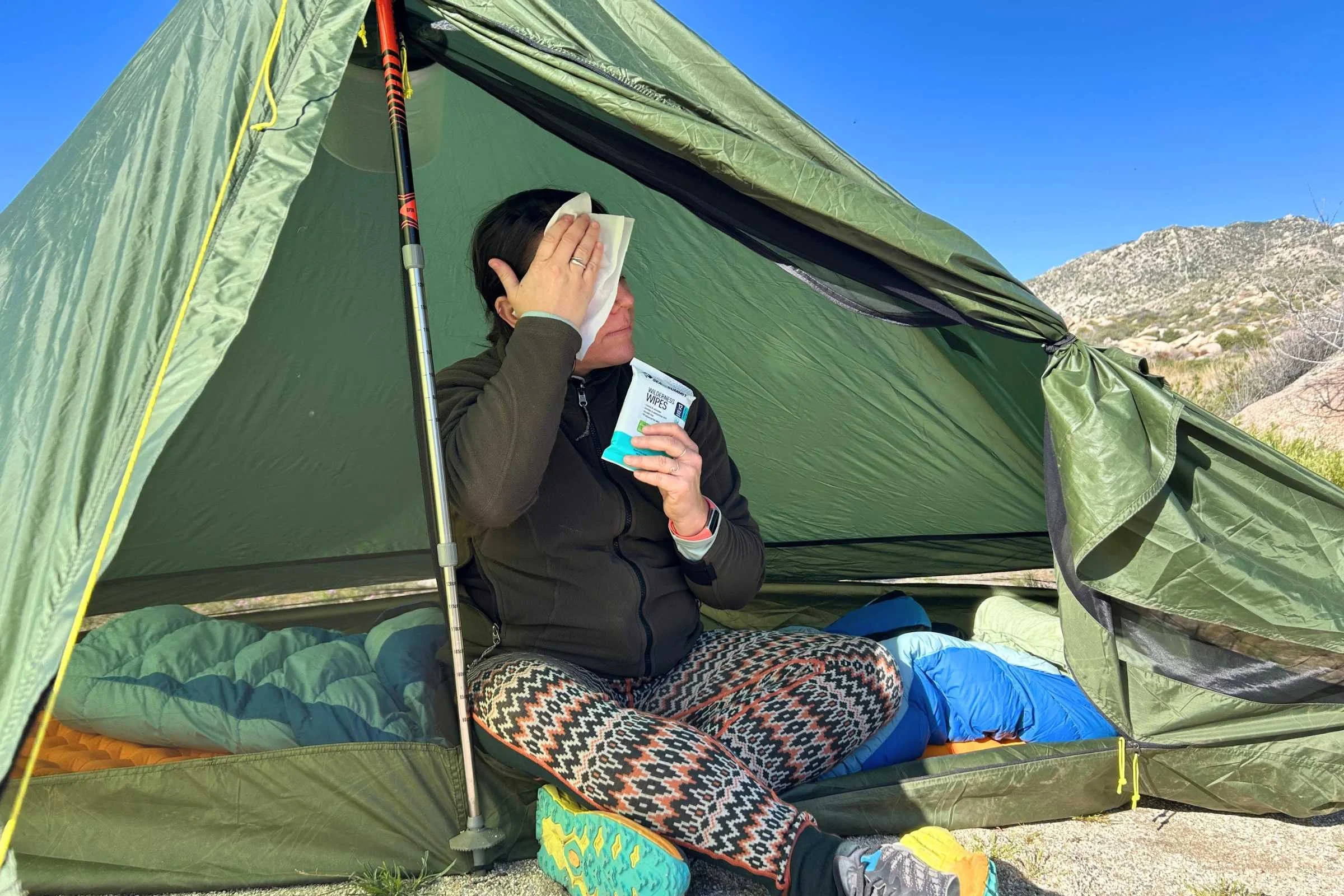 A woman sitting in a green tent with hills in the background uses a Sea to Summit Wilderness Wipe to clean her face