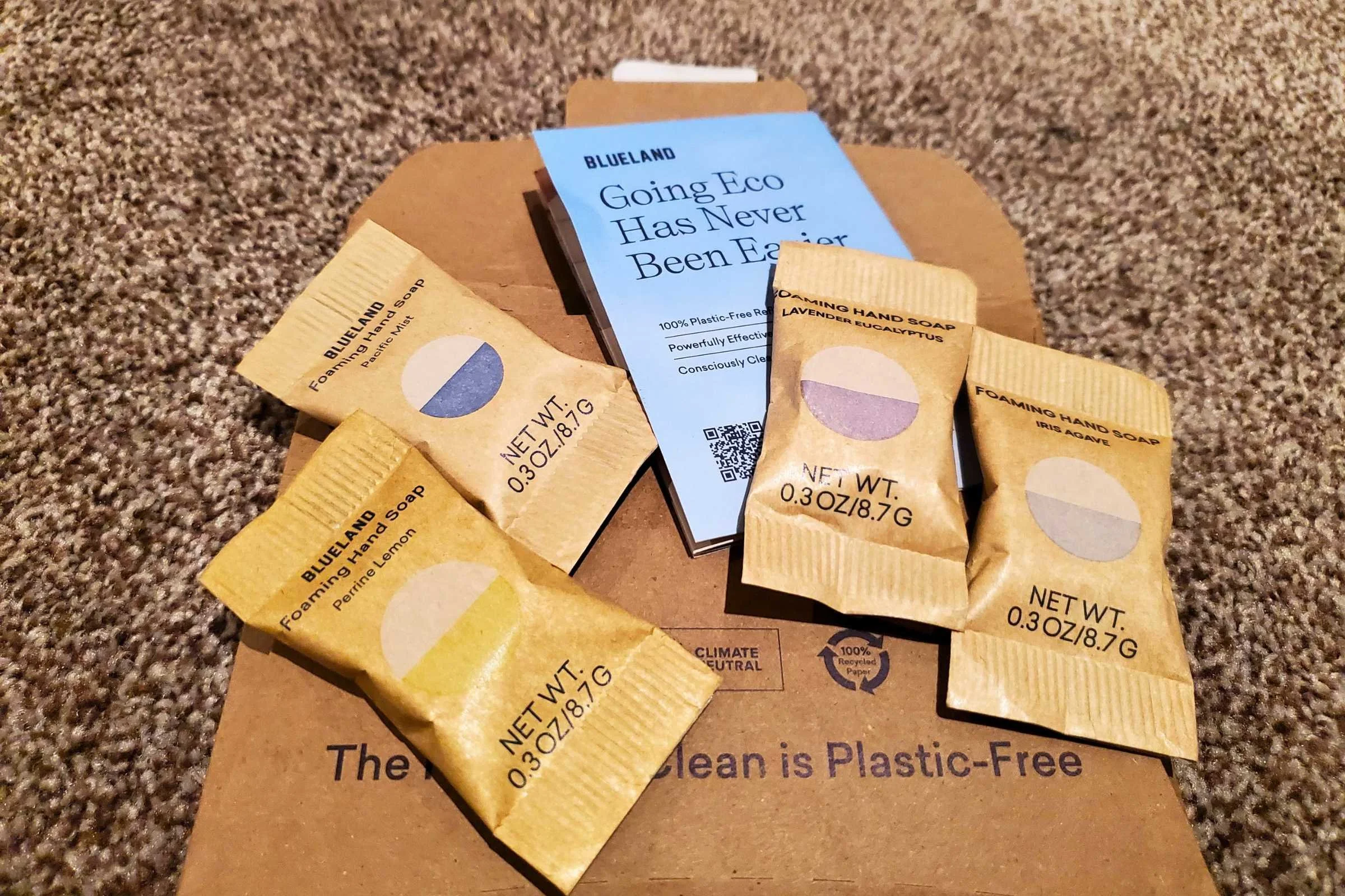A collection of Blueland soap tablets wrapped in tan compostable paper packets sit on their cardboard delivery envelope.