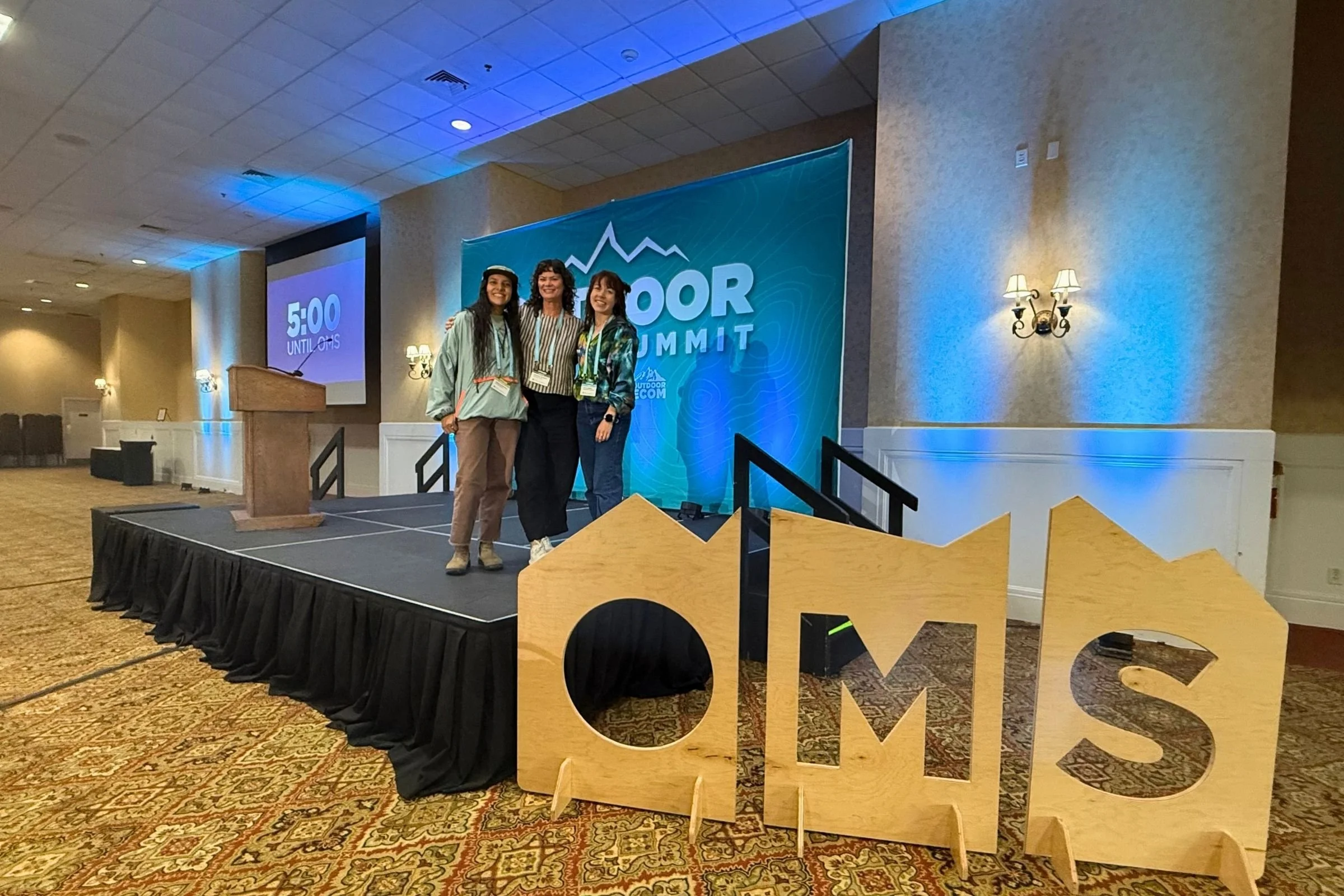 Three women stand on stage in front of a large OMS sign at the 2026 Outdoor Media Summit.