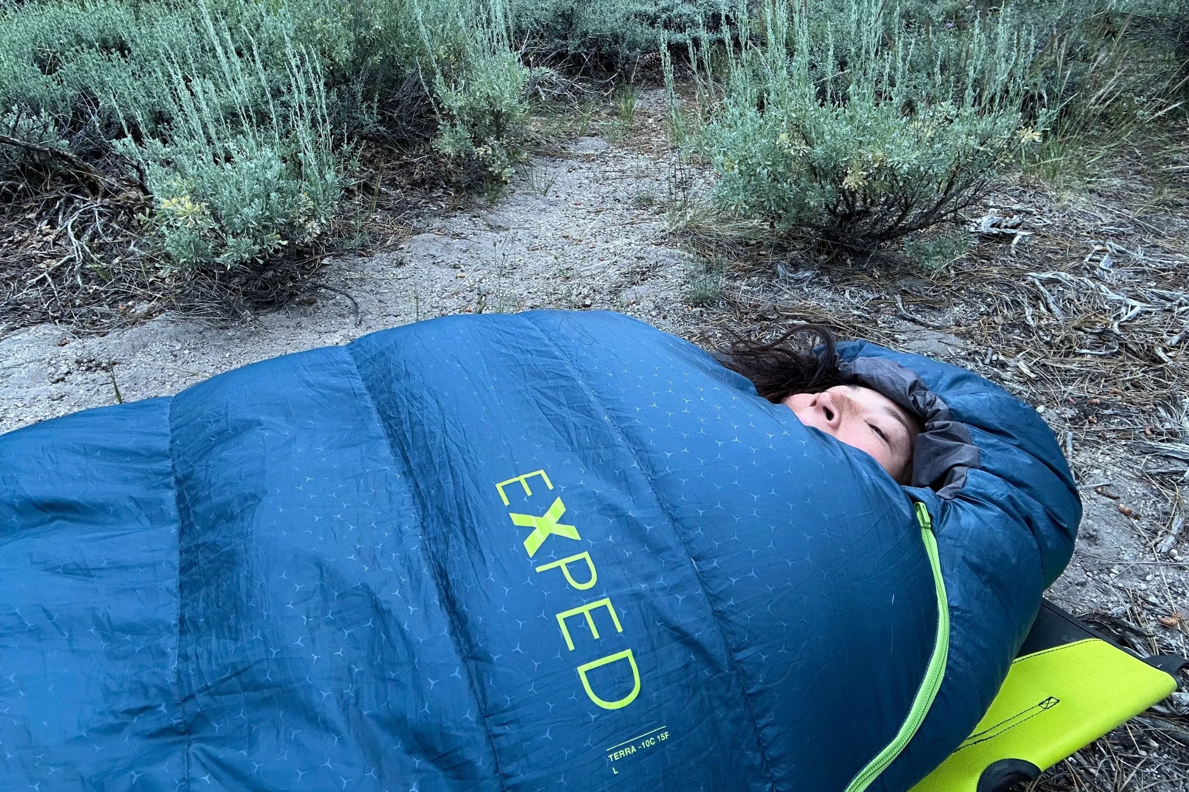 A person lies on the ground inside an Exped Terra Sleeping Bag on top of a camping cot.