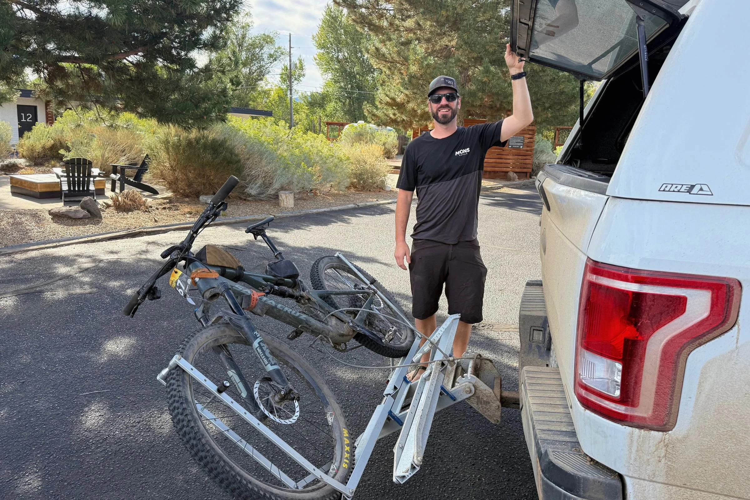 Person standing beside a bicycle in a truck bed, with the 1UP USA Heavy Duty Hitch Bike Rack tilted and loaded.