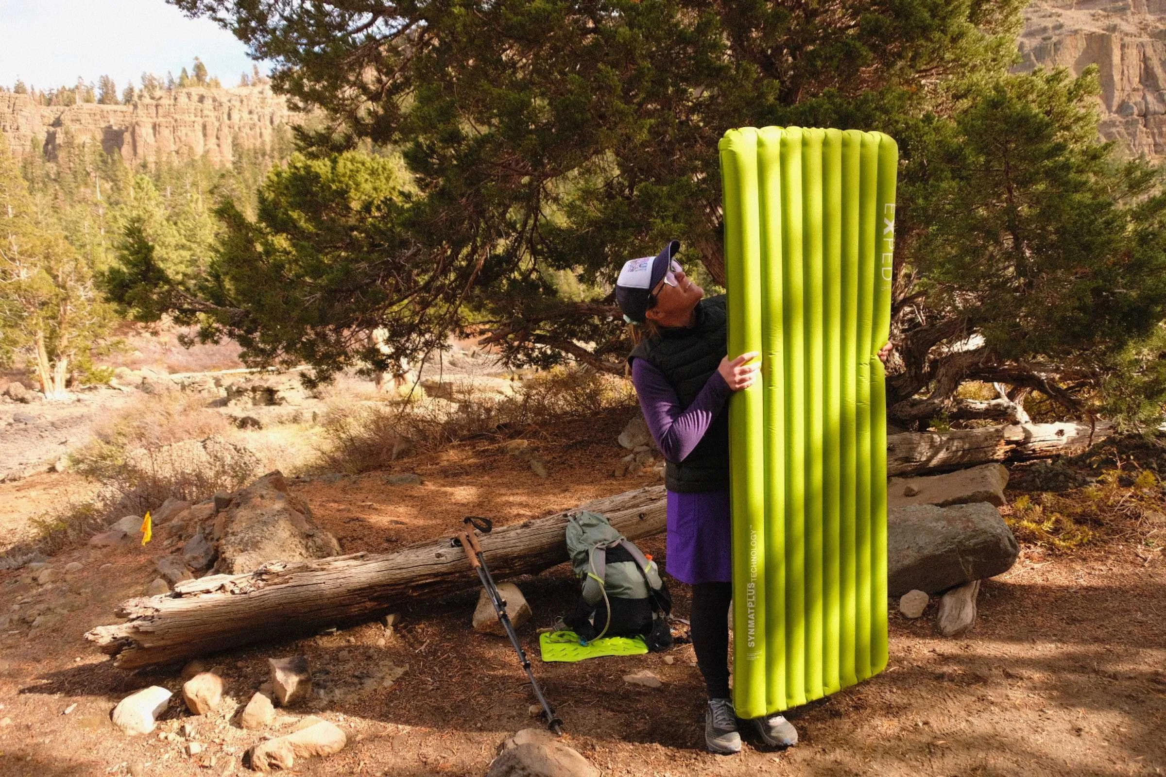 Tester holding EXPED Ultra 6.5R rectangle sleeping pad at campsite surrounded by trees and rocky cliffs