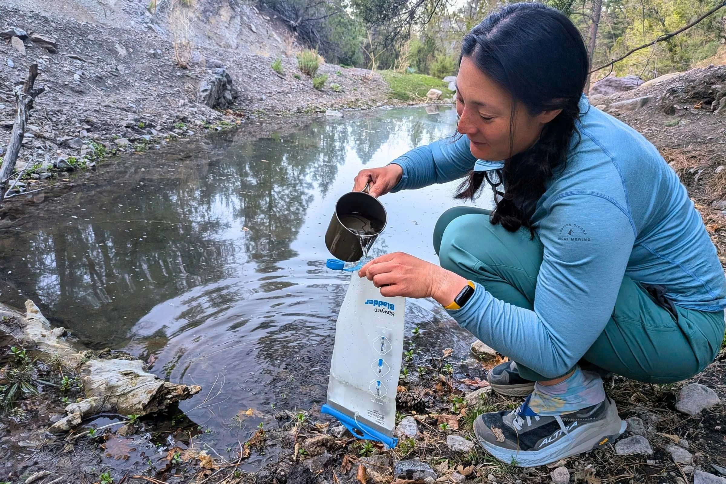 Backpacker squatting by a water source using a backpacking cook pot to fill a Sawyer water bladder