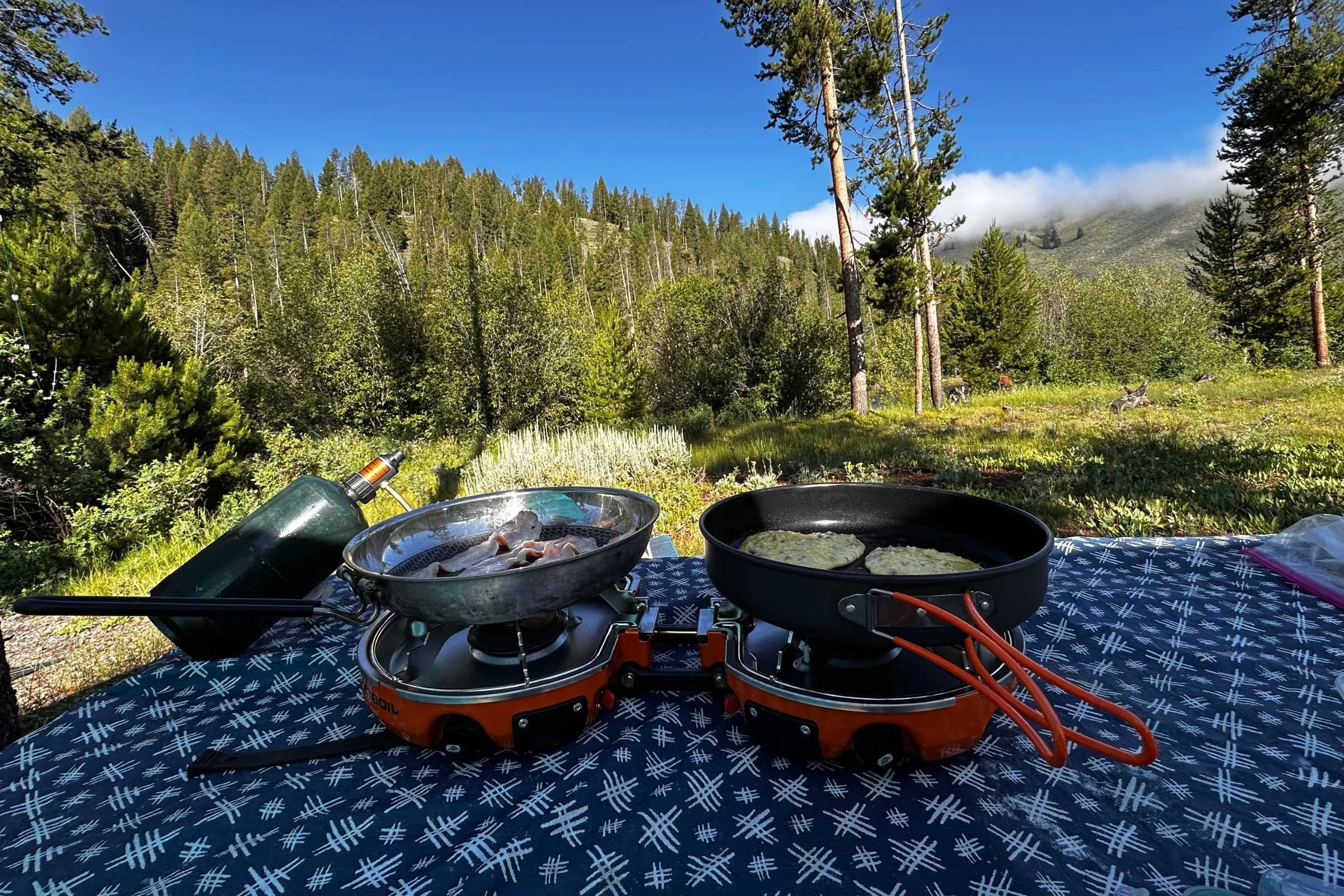 The two burner Jetboil genesis sits on a blue tablecloth with two skillets sitting on each burner. One skillet is cooking pancakes and the other skillet is cooking bacon.