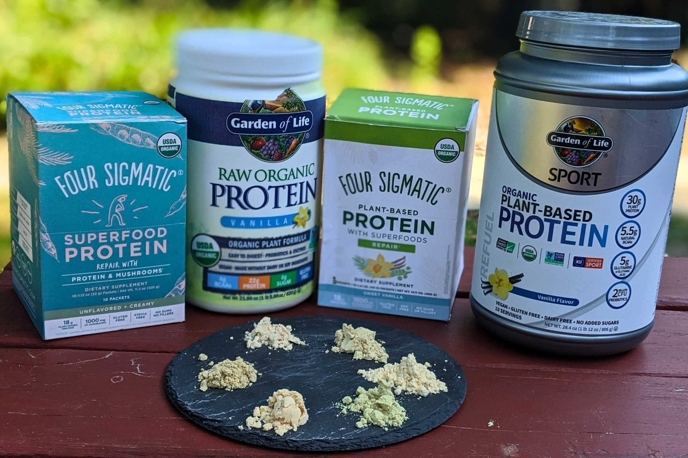   Four protein powder products lined up on a table with sample scoops of each powder in front: Four Sigmatic Superfood Protein, Garden of Life Raw Organic Protein, Four Sigmatic Plant-Based Protein, and Garden of Life Sport Plant-Based Protein.  