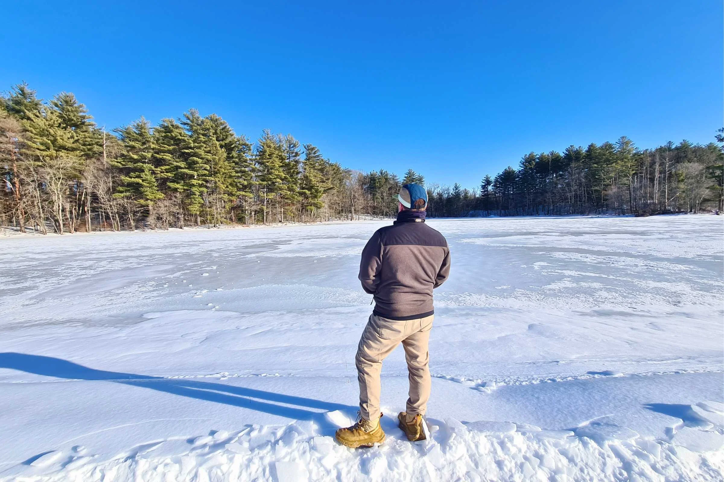   A person standing near a frozen lake wearing the HOKA Kaha 2 Frost GTX hiking boots in winter conditions  