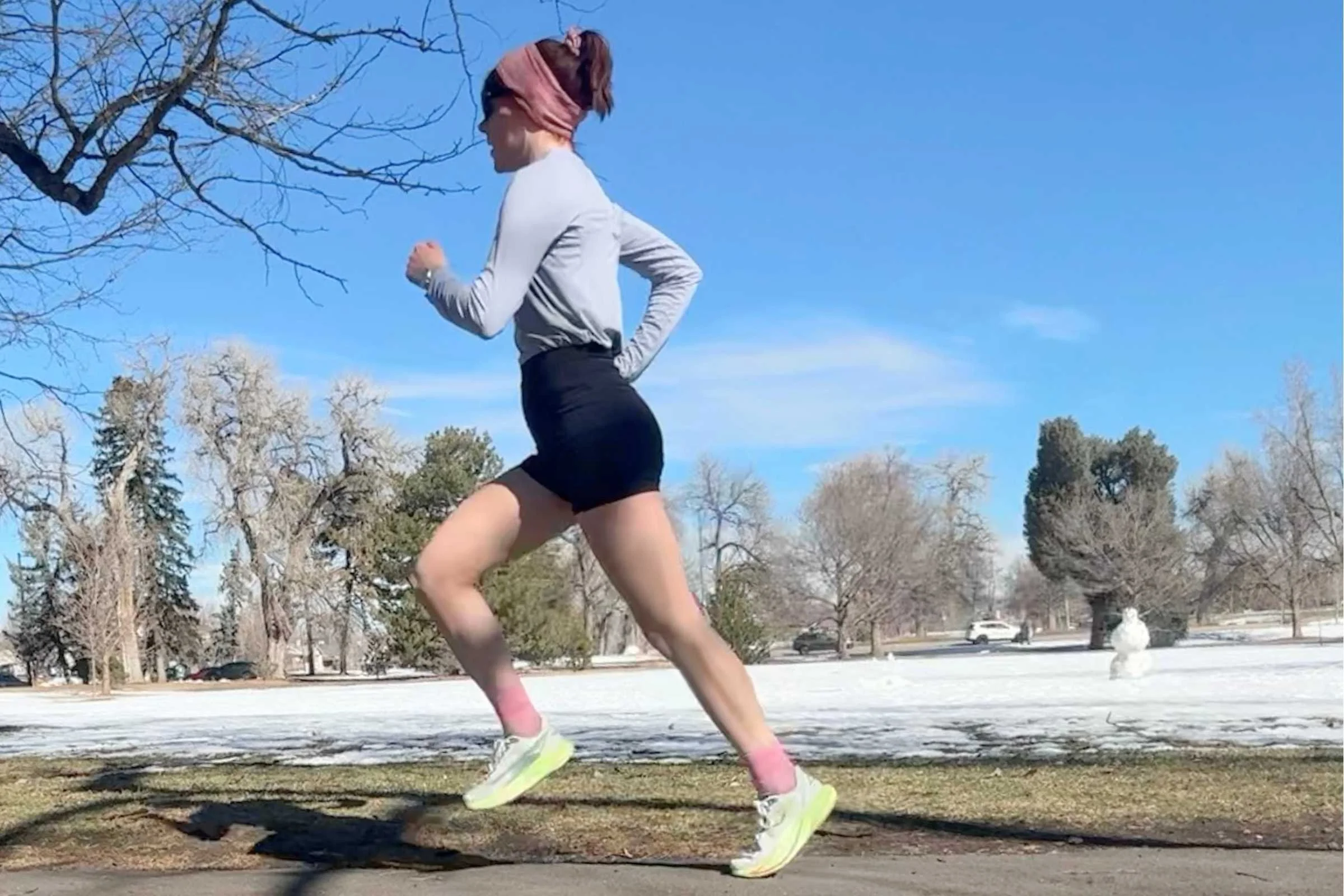   Woman running on a snowy park path in HOKA Mach 7 running shoes under a bright blue sky.  