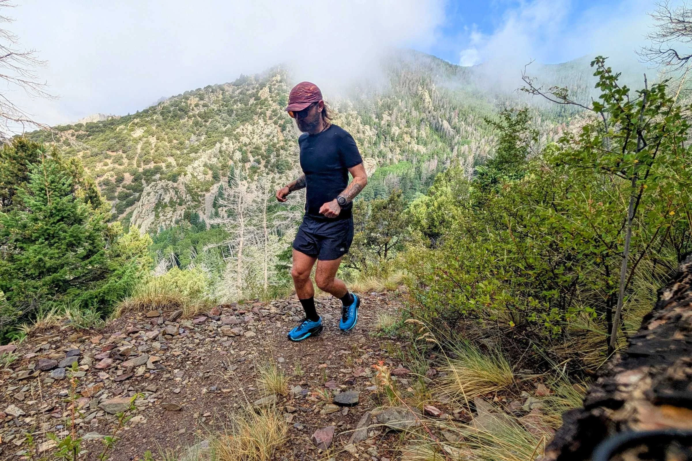   Man running on a mountain trail wearing REI Swiftland shorts with green hills and low clouds in the background.  