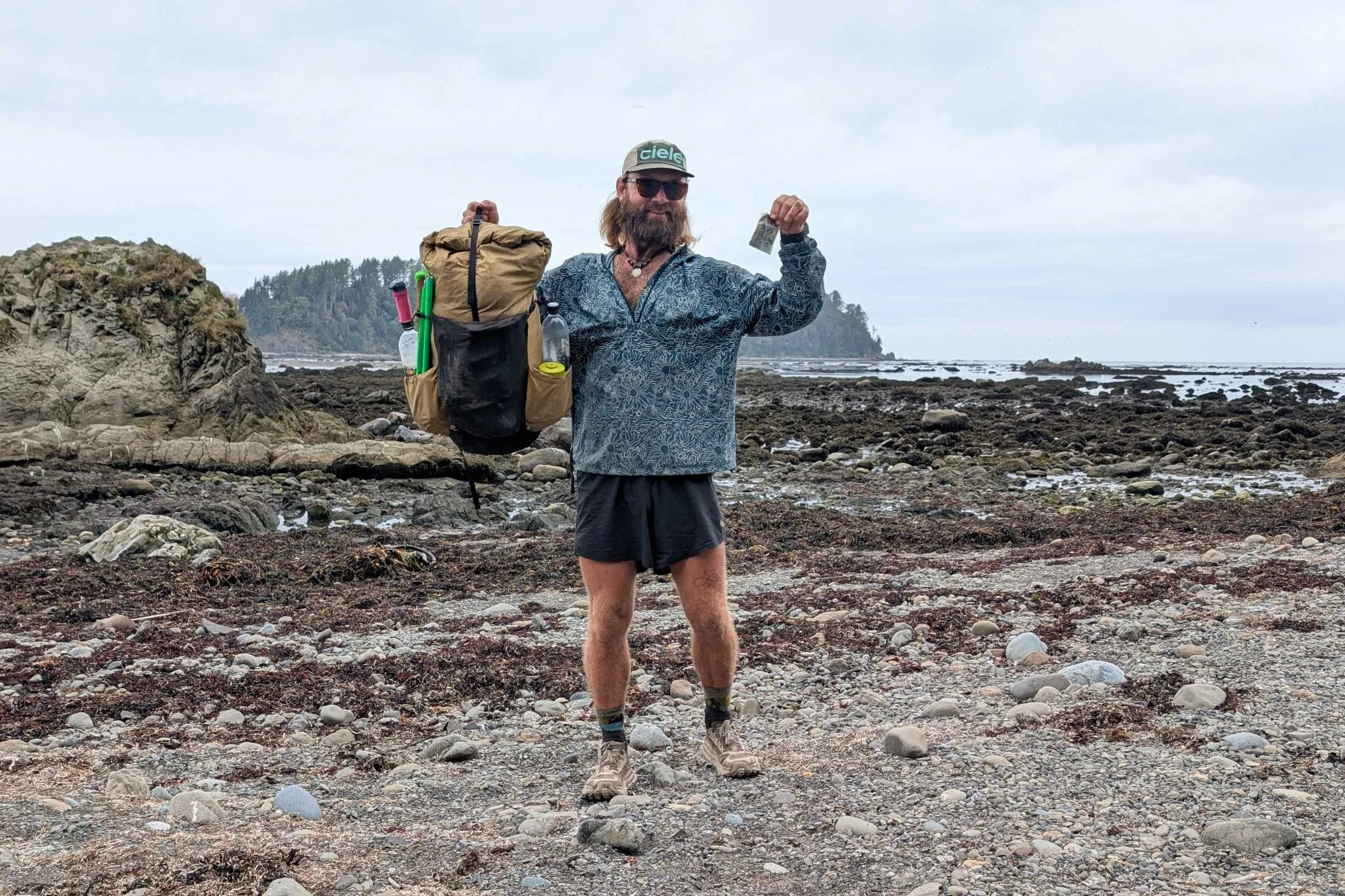   Man walking on rocky coastal terrain wearing REI Co-op Swiftland 5-inch Running Shorts holding up a pack.  