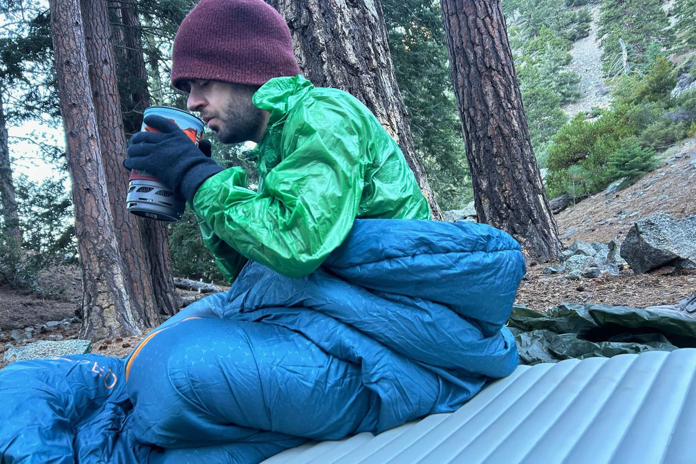   Camper sitting in Exped DeepSleep sleeping bag while drinking hot beverage during cold-weather camping test  