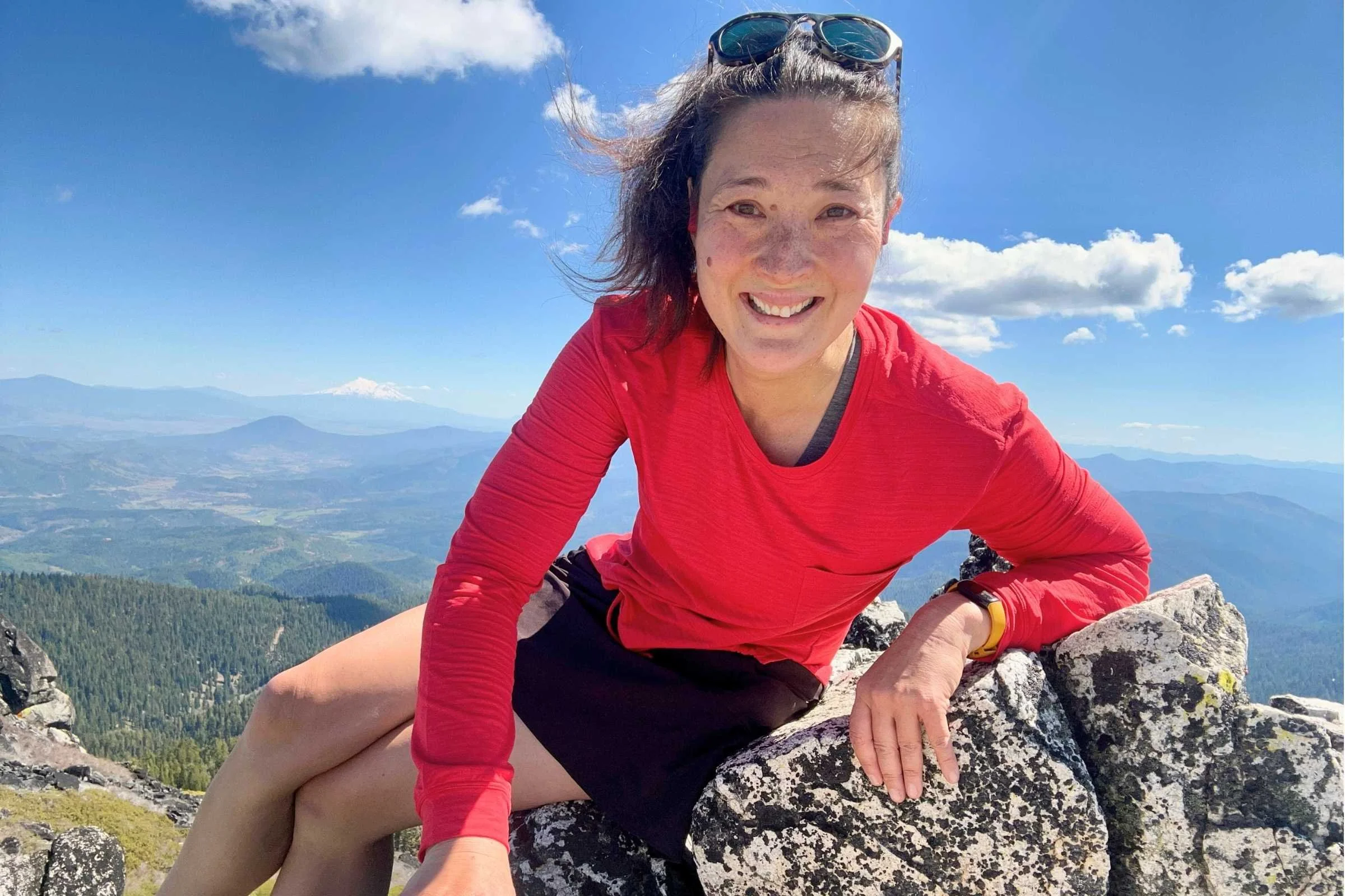   Woman wearing Title Nine Upland Sun Shirt while sitting on rocky summit with mountain views in the background  