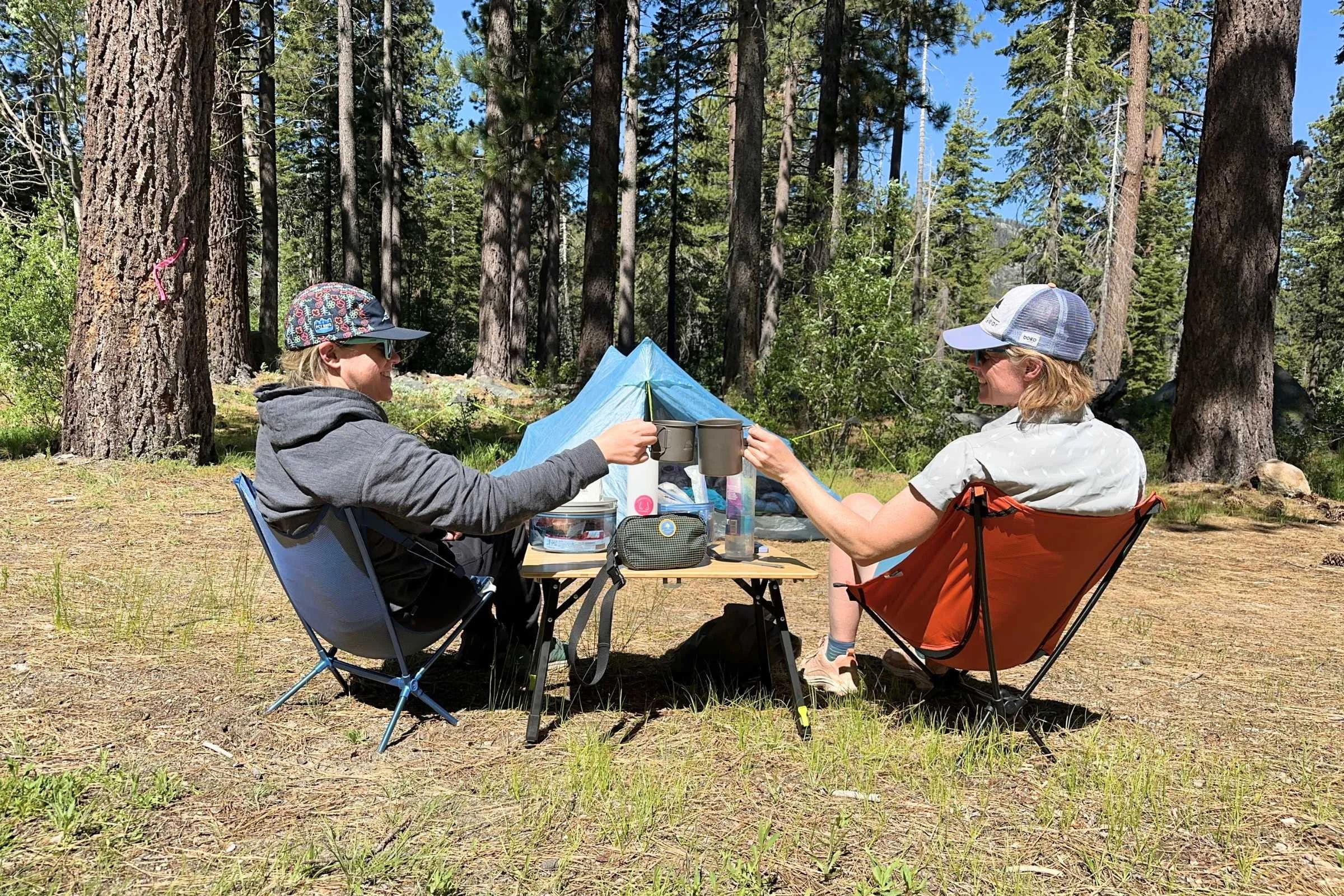   Two campers drinking coffee while sitting at the Zempire Kitpac Pro table set at a lower height.  
