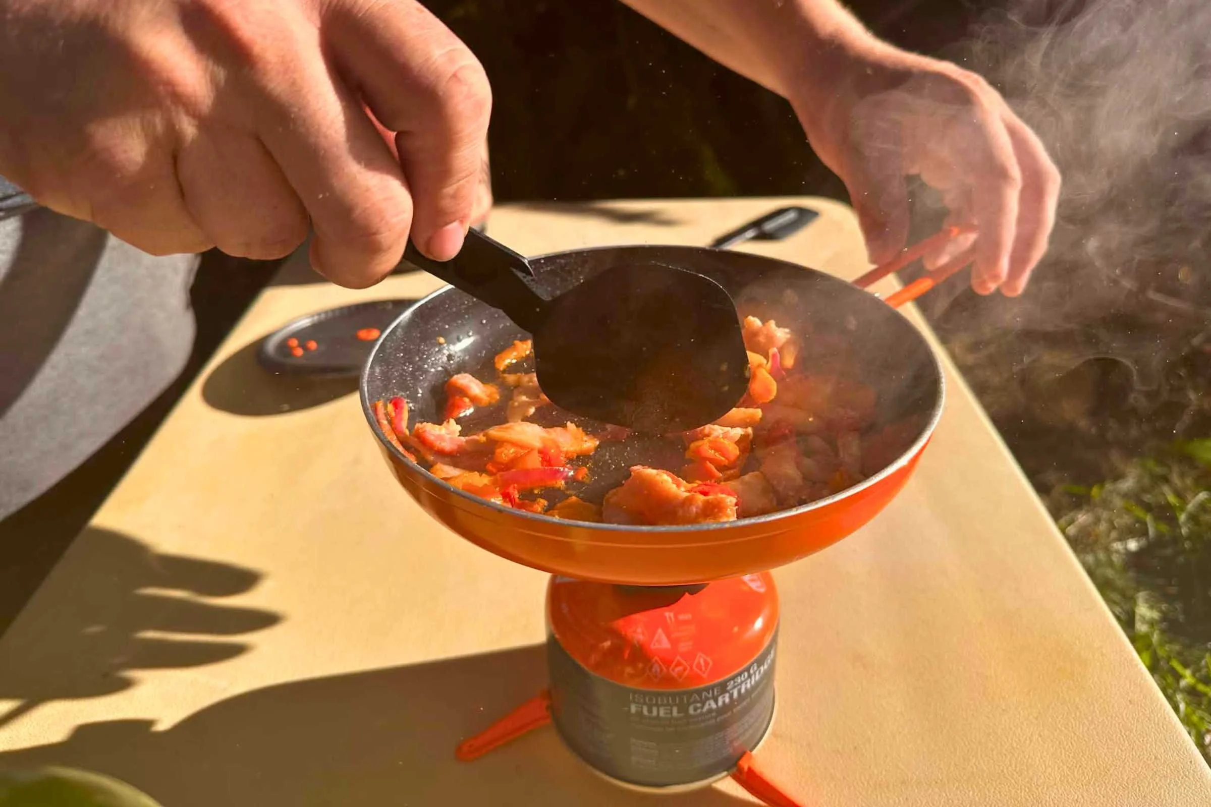   One hand holds the handle of the Jetboil Summit Skillet while another uses the included spatula to stir meat and vegetables as they cook on the TrailCook stove.  