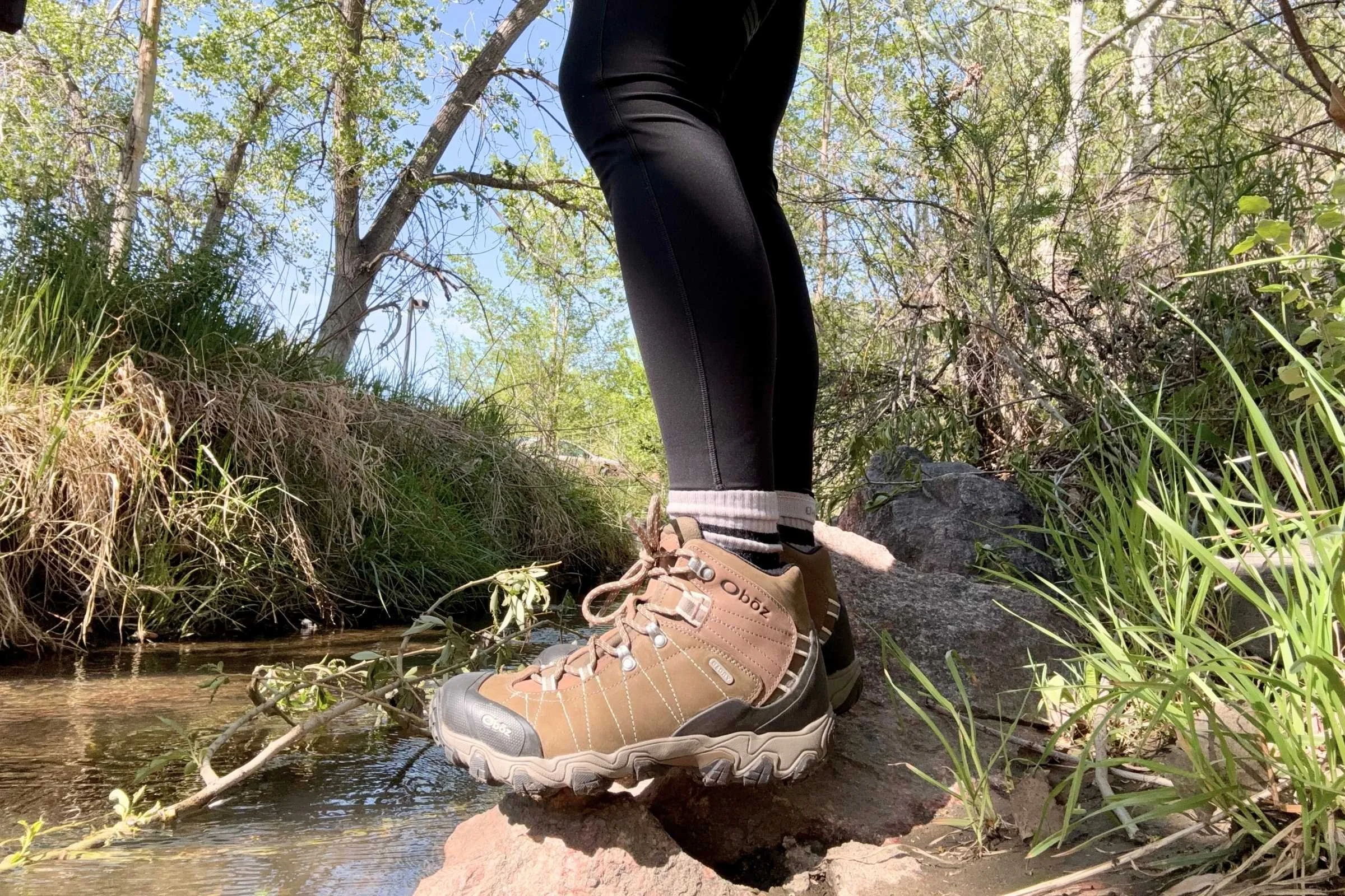   A hiker steps across a rocky stream in the Oboz Bridger Mid Waterproof boots, highlighting their stability and protection on wet terrain.  