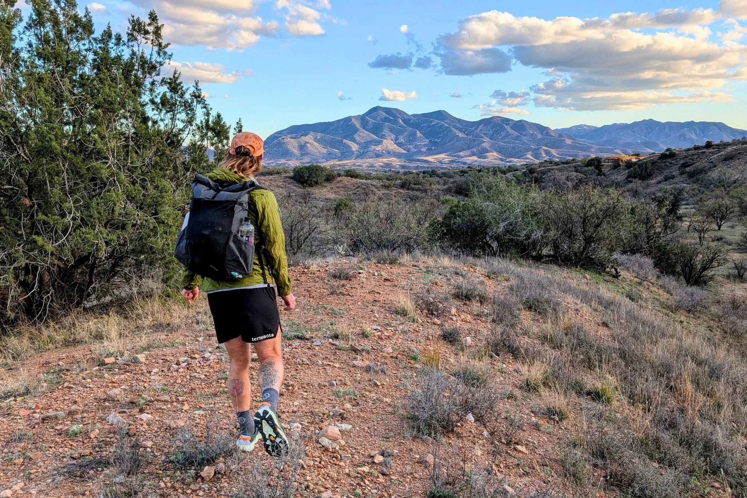   A hiker wearing the Pa’lante V2 with Joey Straps walks along a rocky desert trail with mountains in the background.  