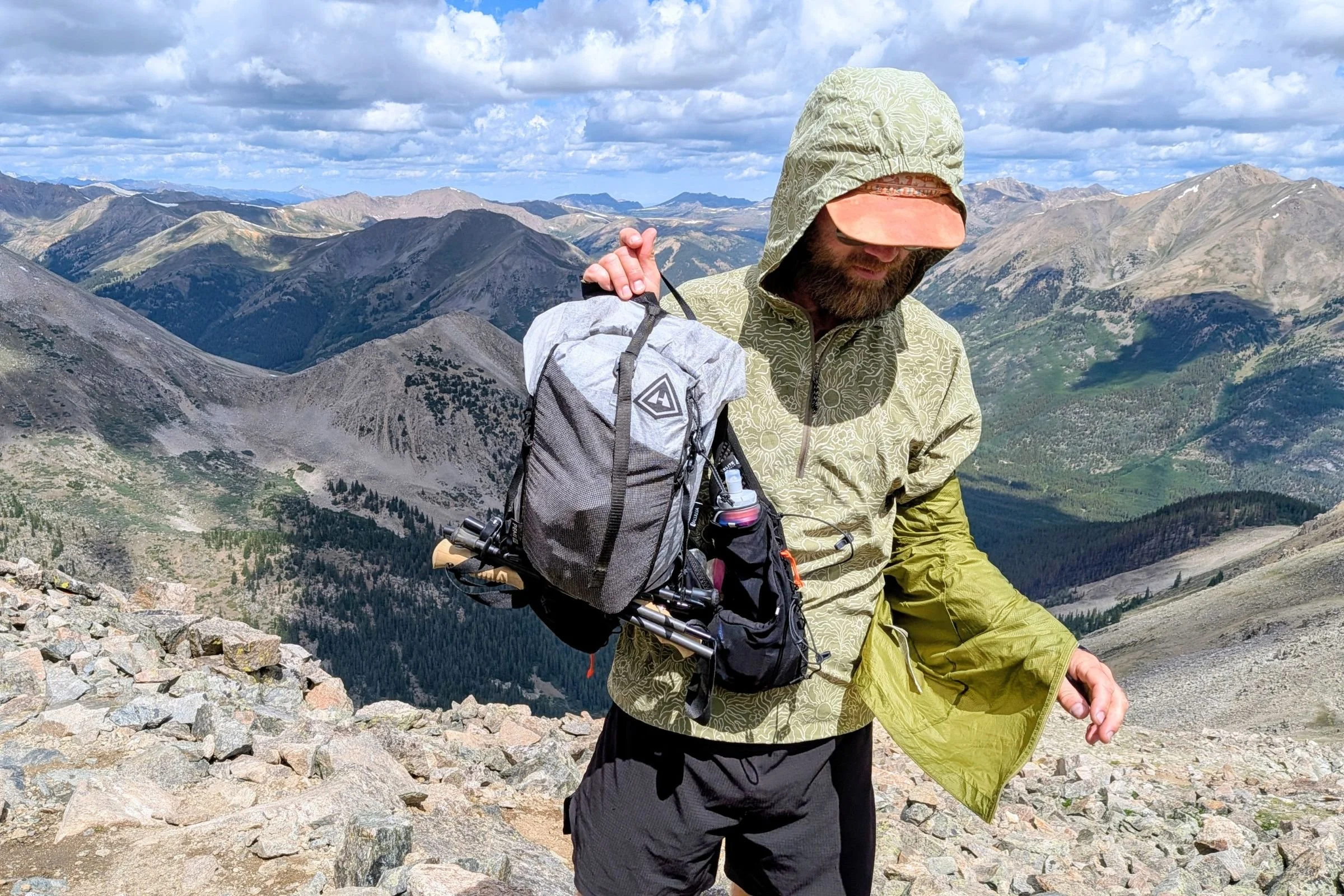   A tester holds the Hyperlite Mountain Gear Pemi 15 against a backdrop of rugged mountain terrain, showing the compact fastpack and its vest-style shoulder straps.  