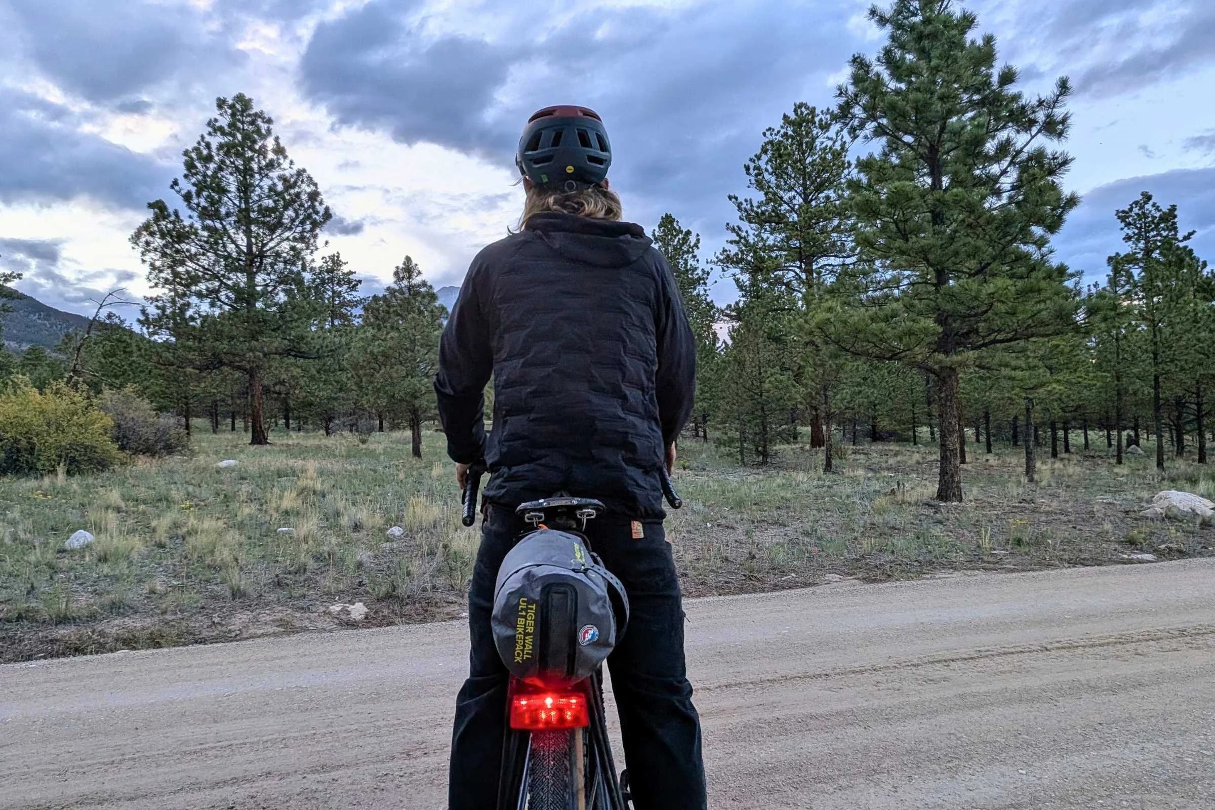   A bikepacker rides a dirt road with the Portland Design Works OxRack Rear Rack mounted above the rear wheel and a tail light secured to the back of the rack.  