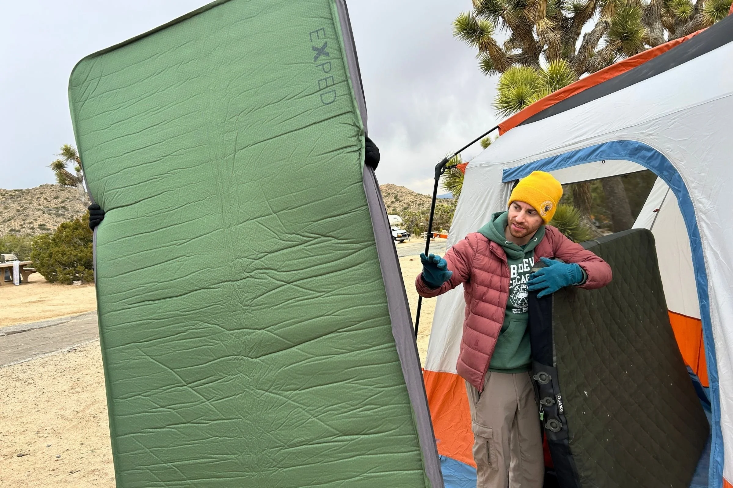   A tester holds the EXPED MegaMat Duo upright outside a tent, showing the mattress’s large dimensions for two campers.  