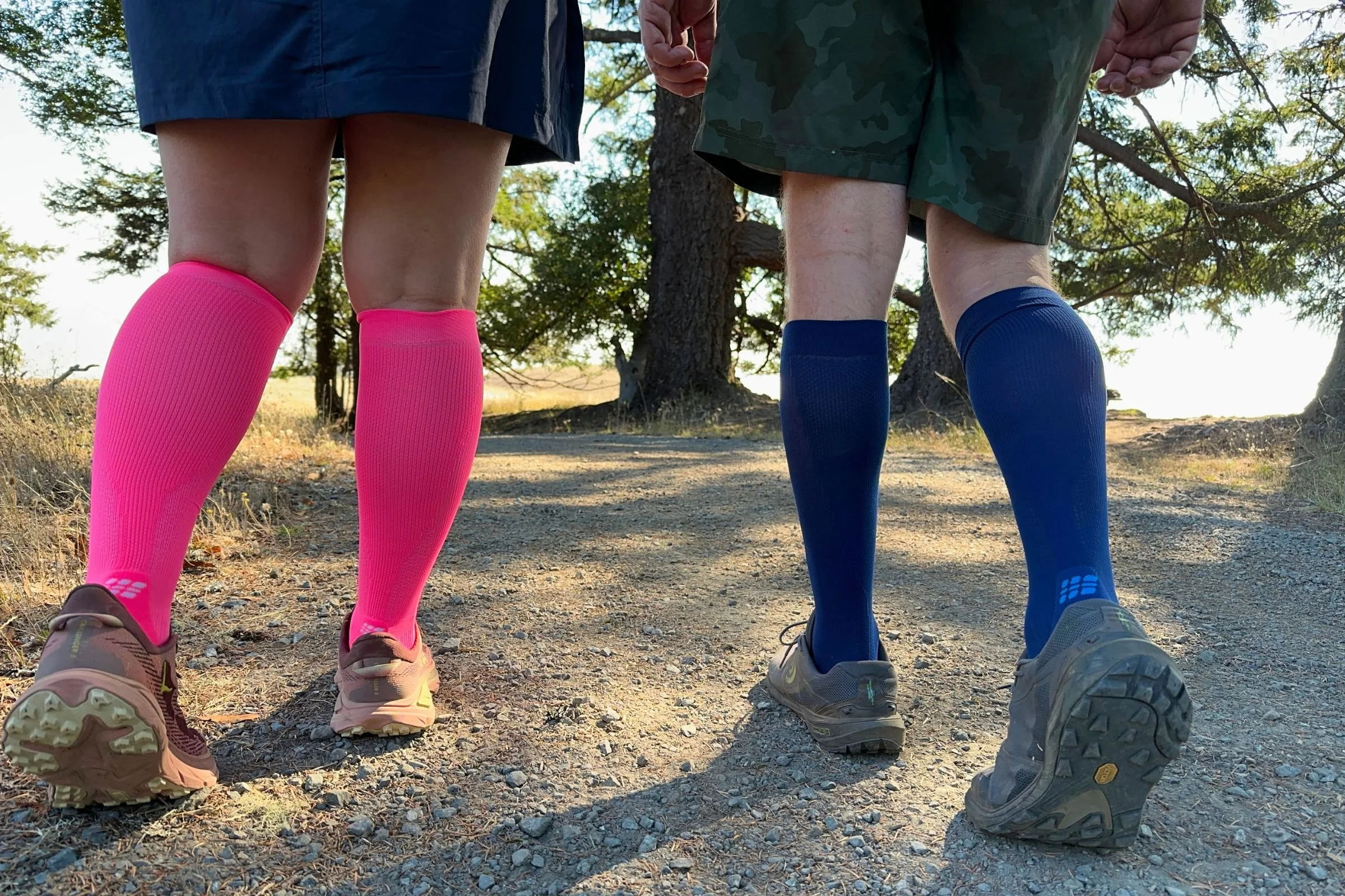   Two runners wear the CEP Core Run Tall Compression Socks 5.0 in bright pink and navy blue on a dirt trail, with graduated 20–30 mmHg compression designed to provide comfortable support during runs and recovery.  